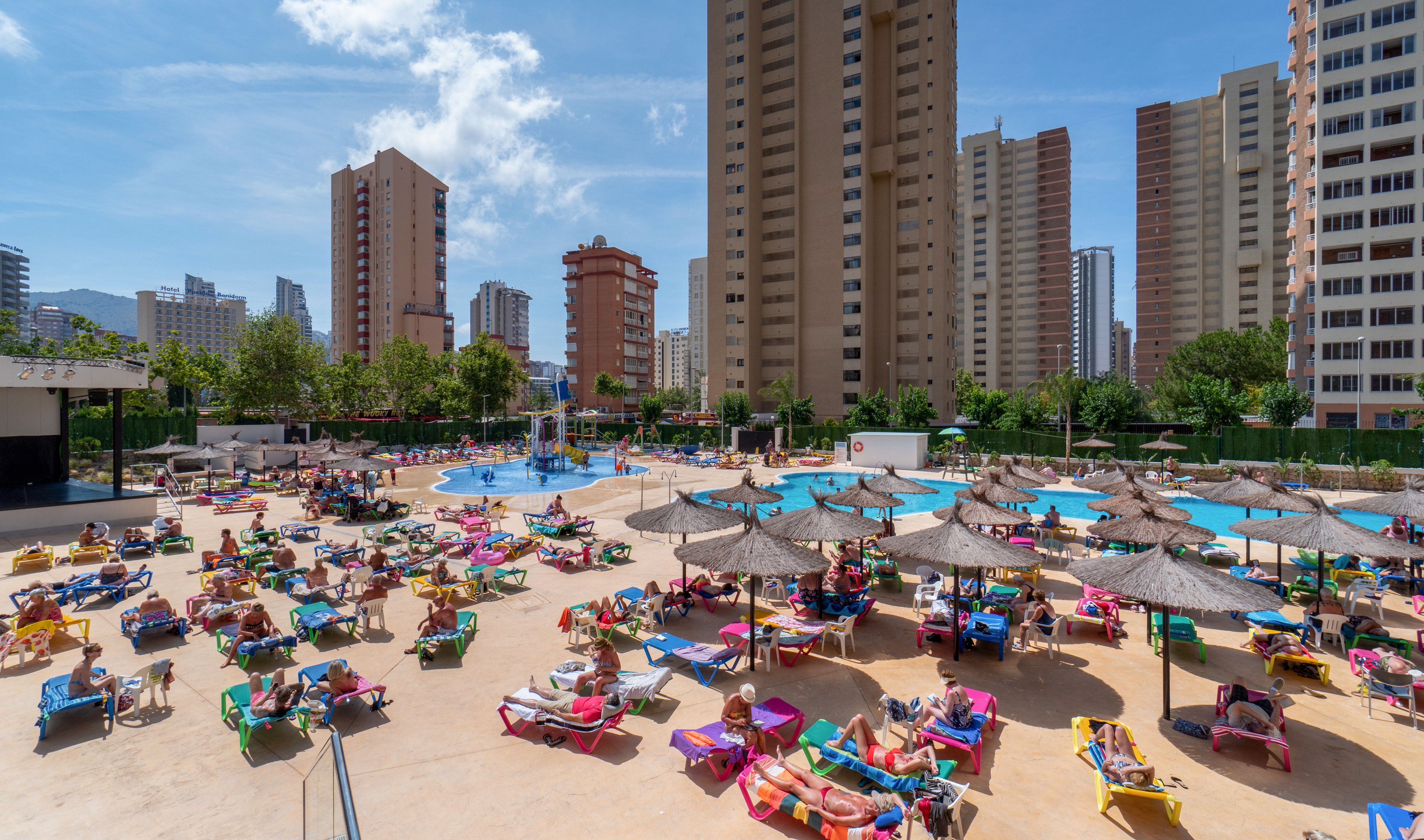 A large outdoor resort pool area is filled with colorful sun loungers and umbrellas under bright sunshine. Multiple swimming pools and thatched parasols are surrounded by vacationers relaxing and sunbathing. Tall residential and hotel towers rise in the background, framing the urban holiday setting. The clear blue sky and vibrant colors create a lively, summery atmosphere.