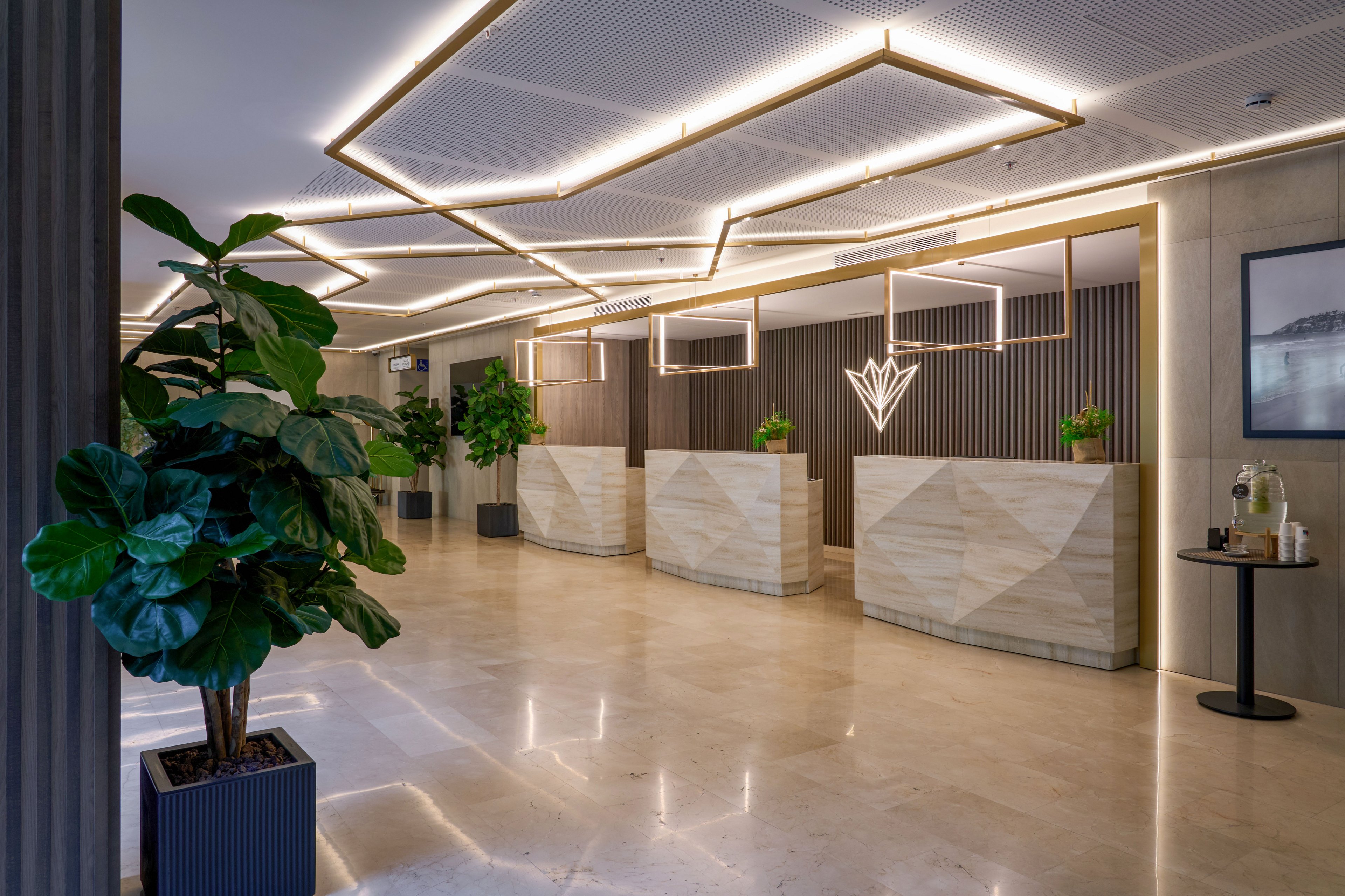 Contemporary hotel reception area featuring three marble-fronted check-in desks. The space is illuminated by geometric ceiling lights that create a warm, inviting atmosphere. Large potted plants and minimalist decor add a touch of greenery and sophistication. A small side table with refreshments and wall art completes the stylish lobby setting.