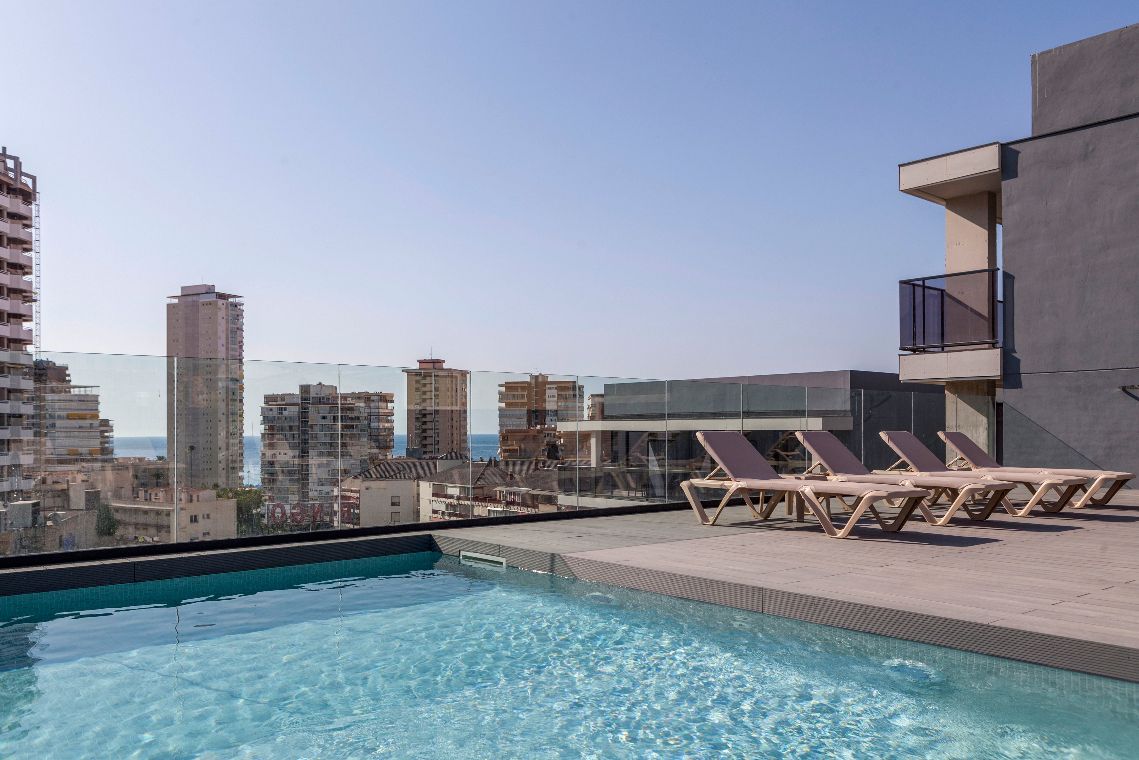 A modern rooftop swimming pool sits beside a row of sun loungers on a spacious terrace. Glass railings frame the edge, revealing a dense city skyline of mid-rise buildings. In the distance, the sea is visible under a clear blue sky, adding a coastal feel to the urban setting. The overall scene suggests a contemporary, relaxing outdoor leisure space.