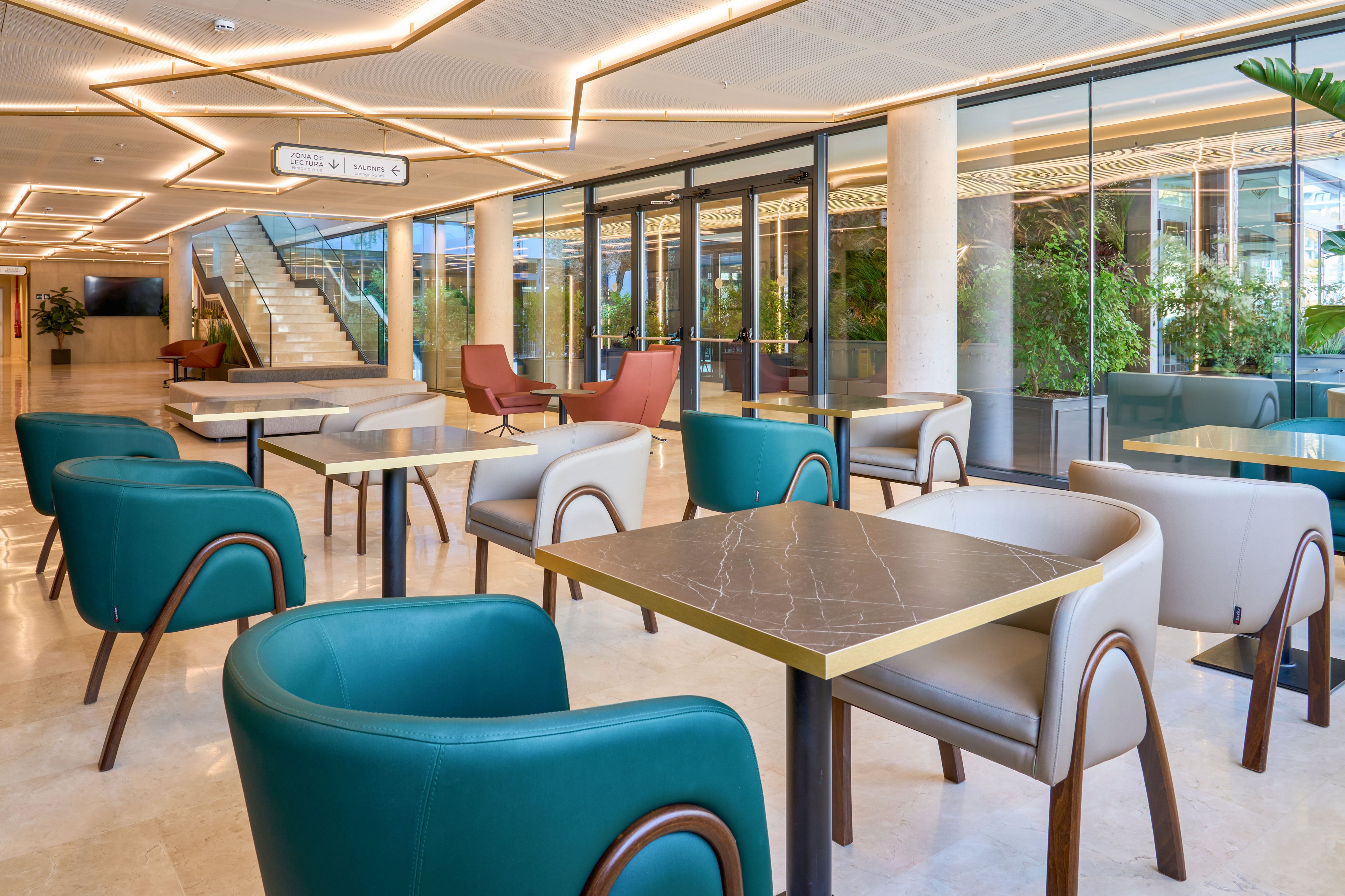 A contemporary lobby lounge features square tables surrounded by teal and beige upholstered armchairs. Large floor-to-ceiling glass windows reveal lush indoor greenery and an upper level beyond. Warm ceiling lighting with geometric lines creates an inviting atmosphere. A staircase and corridor signage in the background suggest a public or corporate building setting.
