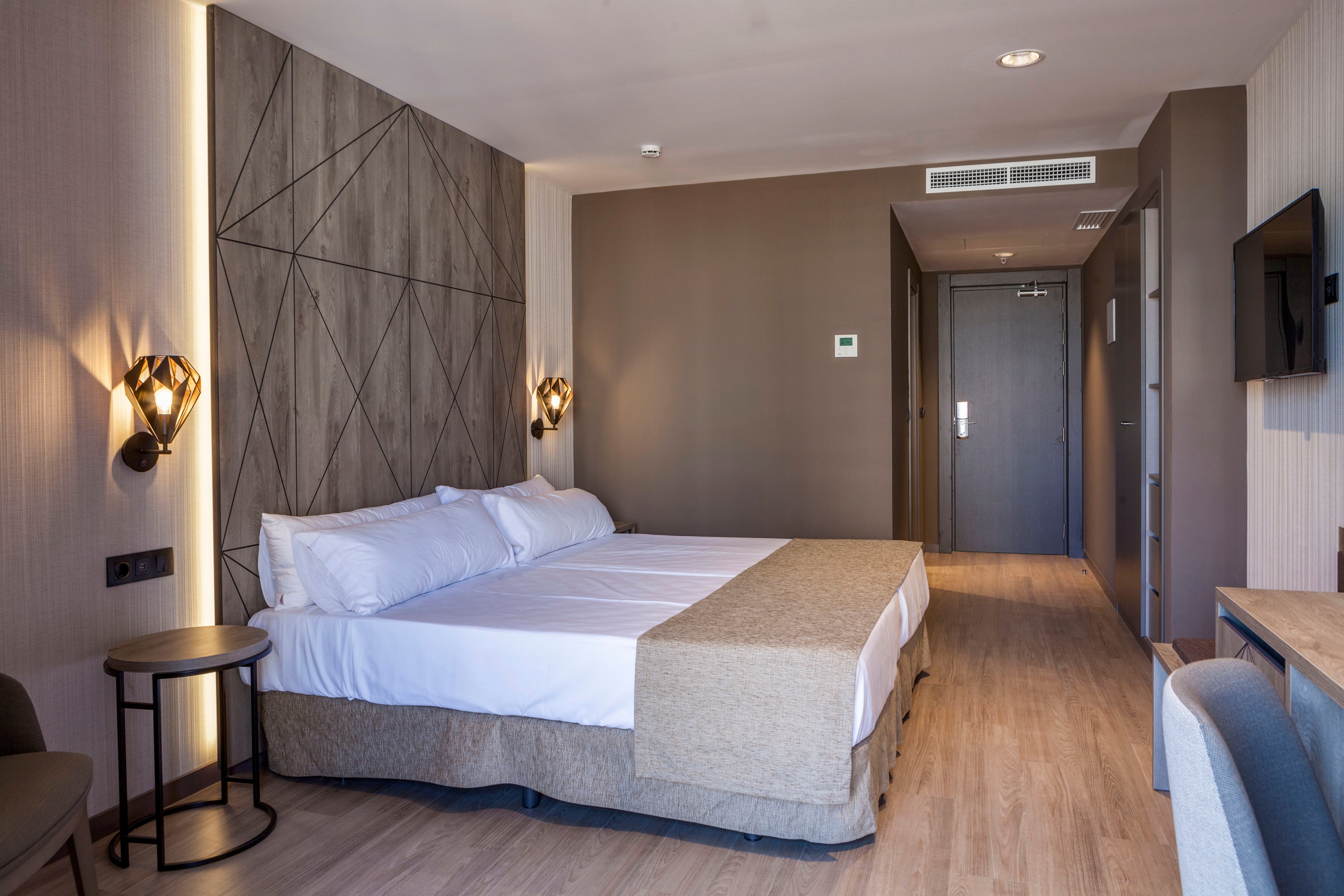 Contemporary hotel room interior featuring a large double bed with white linens and beige bed runner. The space includes wooden flooring, geometric accent wall paneling, and warm wall sconces on either side of the bed. A flat-screen TV is mounted on the wall opposite the bed, with a desk and chair visible in the foreground. The entry door, air conditioning vent, and thermostat are seen at the back of the room, emphasizing a clean, minimalist design.