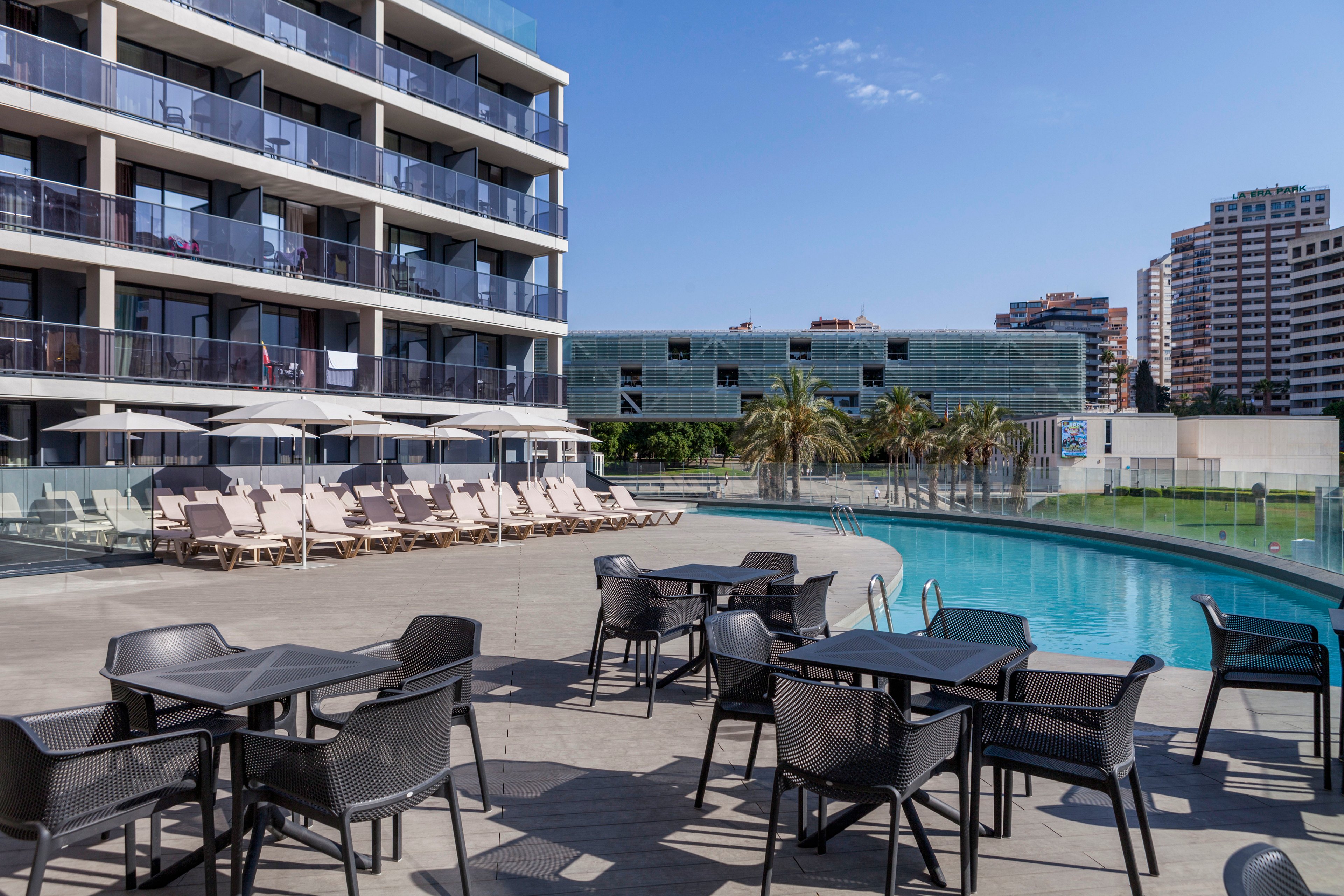 A contemporary hotel terrace features black outdoor tables and chairs beside a curved swimming pool. Sun loungers with umbrellas line the deck in front of a multi-story building with glass balconies. In the background, additional urban buildings and palm trees suggest a city resort setting. The clear blue sky and calm water create a relaxed, vacation atmosphere.