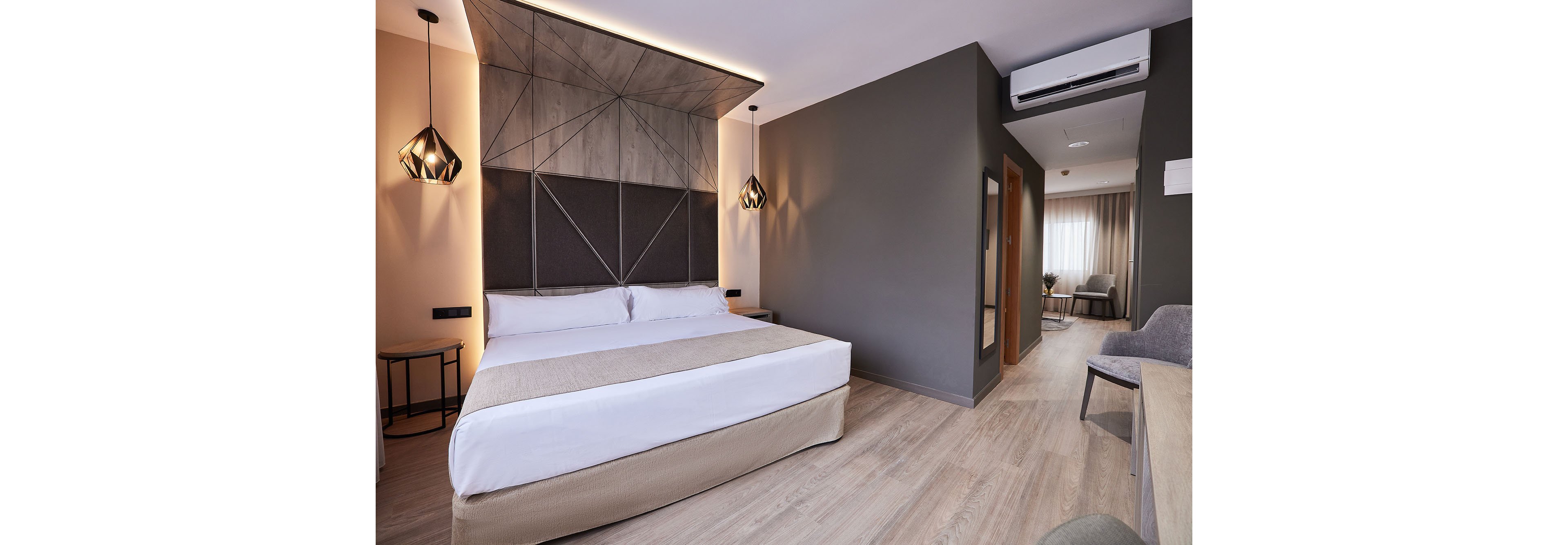 Contemporary hotel room featuring a large double bed with neutral bedding and geometric headboard. The space includes pendant lights, a small side table, and a desk with chair along the wall. Warm wood flooring and soft beige and gray tones create a calm, minimalist atmosphere. A doorway leads to a separate sitting area with armchairs and a window with sheer curtains.
