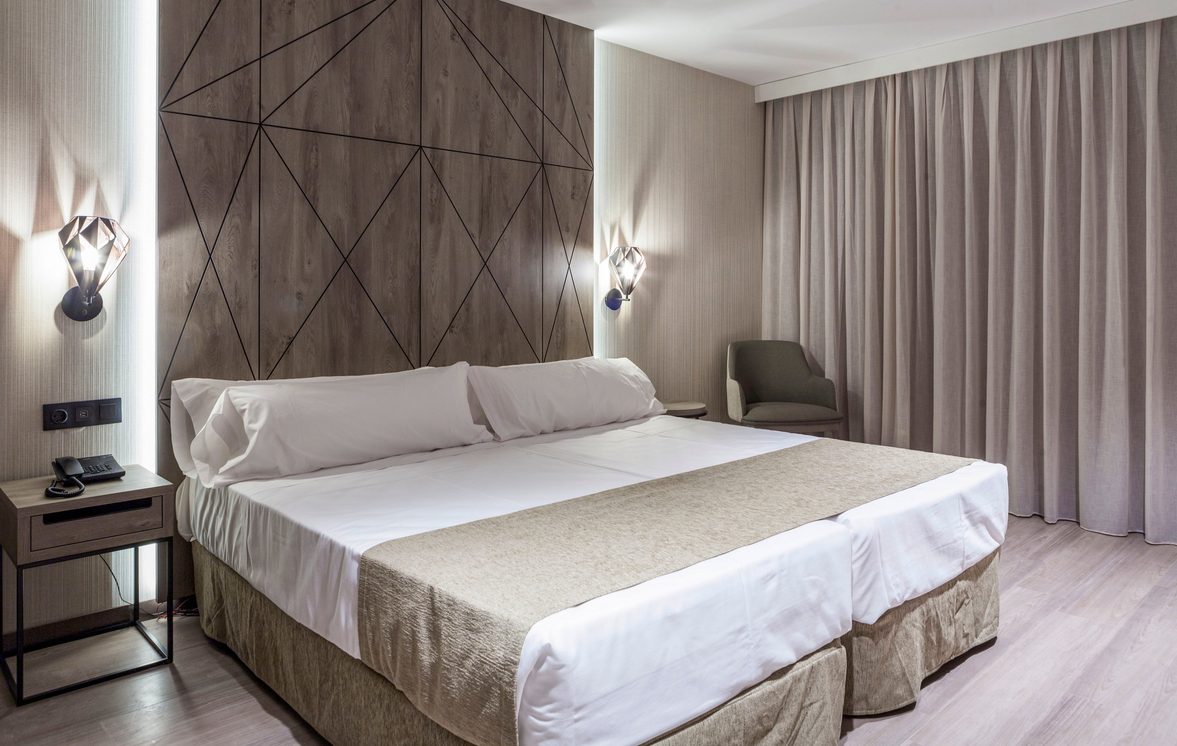 A contemporary hotel room features a large double bed with light beige bedding and white pillows. Geometric wall paneling and two decorative wall sconces frame the headboard, adding a stylish design element. A bedside table with a phone and notepad sits to the left, while a cushioned armchair is placed near floor-length curtains on the right. The overall space appears clean, minimal, and softly lit, creating a calm and inviting atmosphere.