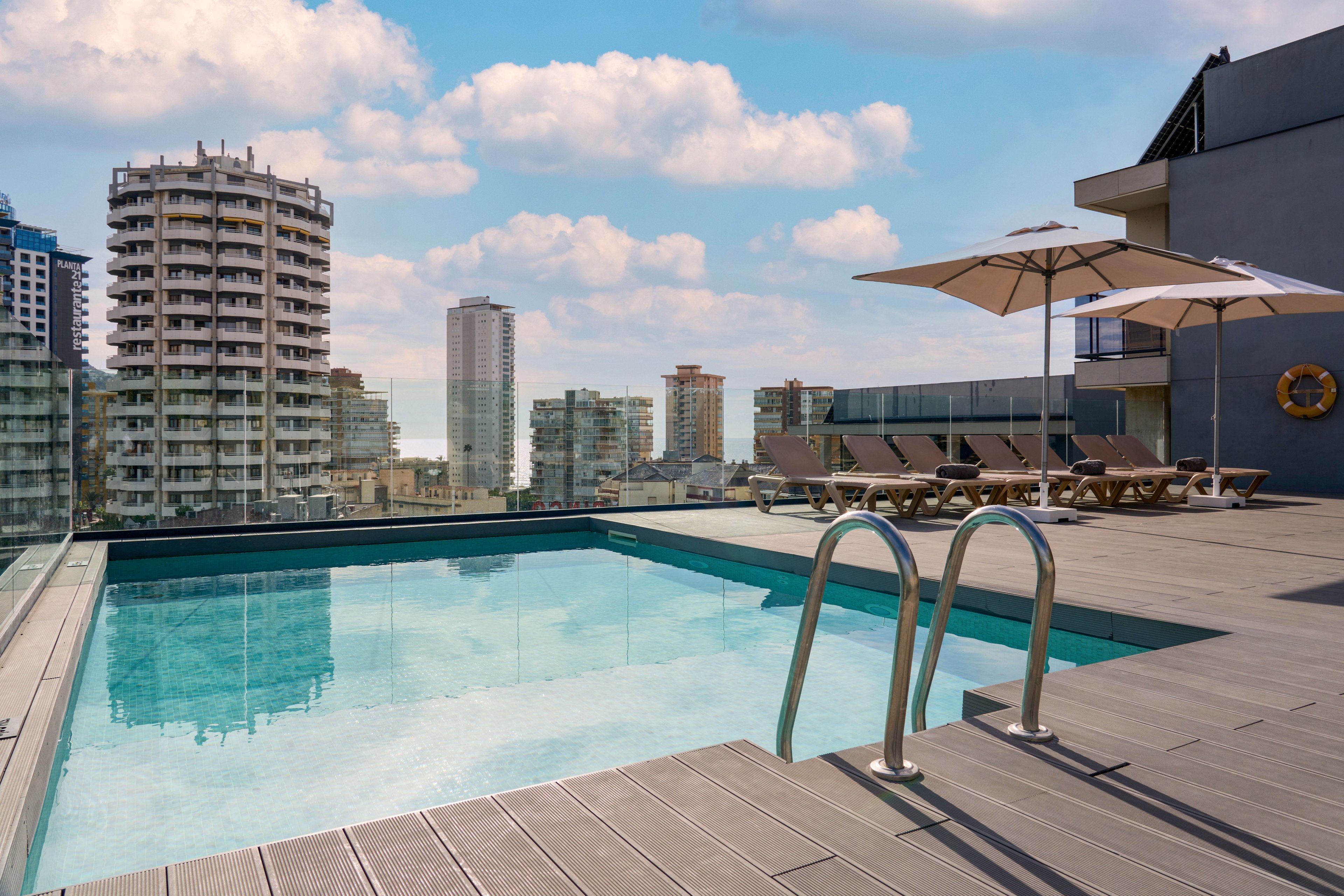 A modern rooftop swimming pool sits atop a building with a metal ladder leading into the clear water. Sun loungers and large patio umbrellas line the deck, suggesting a relaxing leisure area. Surrounding high-rise residential and office towers frame the urban skyline under a partly cloudy blue sky. The scene conveys a contemporary, upscale outdoor environment ideal for relaxation and city views.