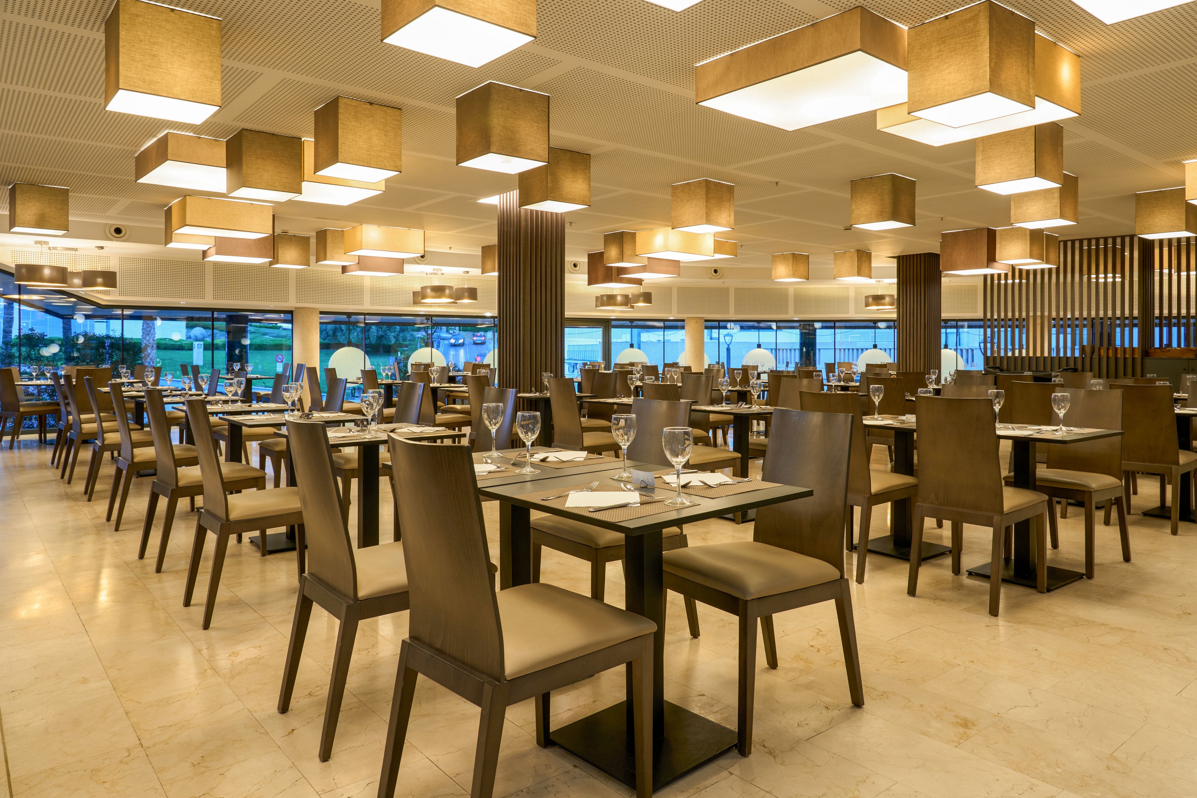 A spacious contemporary restaurant dining area is shown with neatly arranged tables and chairs. Each table is set with glassware, plates, and cutlery, ready for guests. Large windows line one side, revealing an outdoor view and natural light. Warm ceiling fixtures and neutral tones create an inviting, elegant atmosphere.