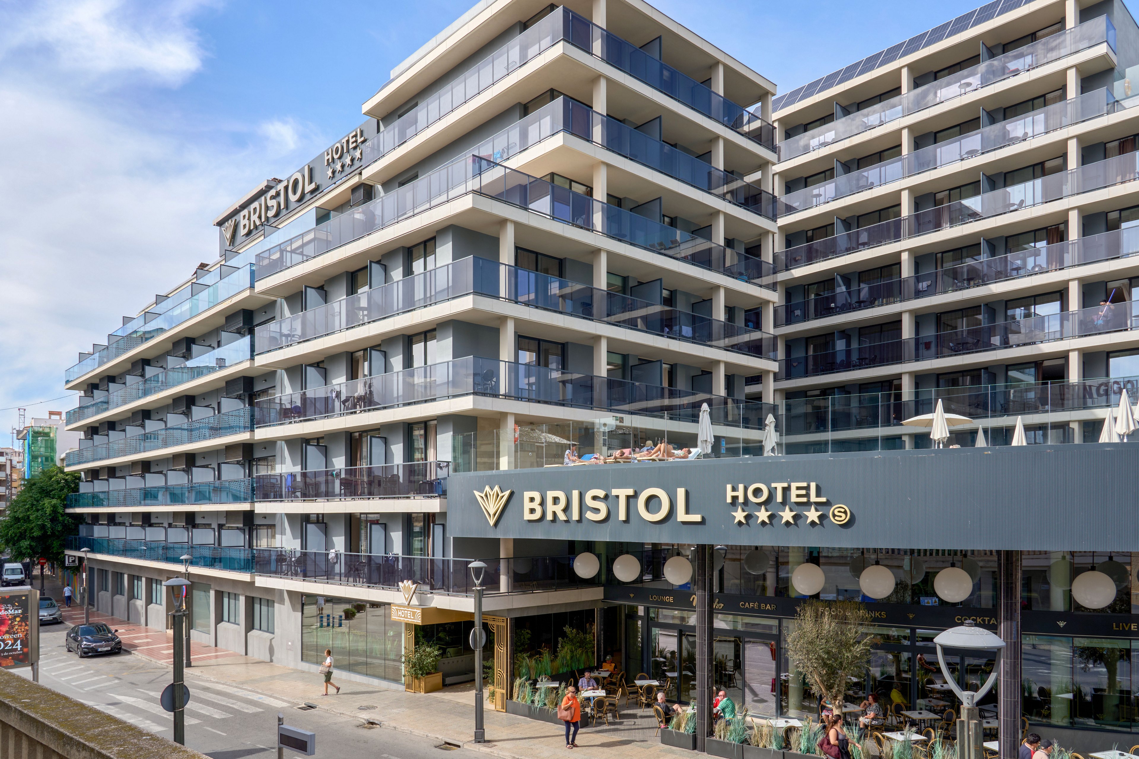 The image shows the contemporary multi-story Bristol Hotel building with numerous glass-fronted balconies. The hotel facade features a prominent sign reading "BRISTOL HOTEL" along with four stars, indicating a four-star rating. People walk along the street and sit at the ground-floor terrace, adding urban activity to the scene. The overall atmosphere is bright and modern under a clear blue sky.