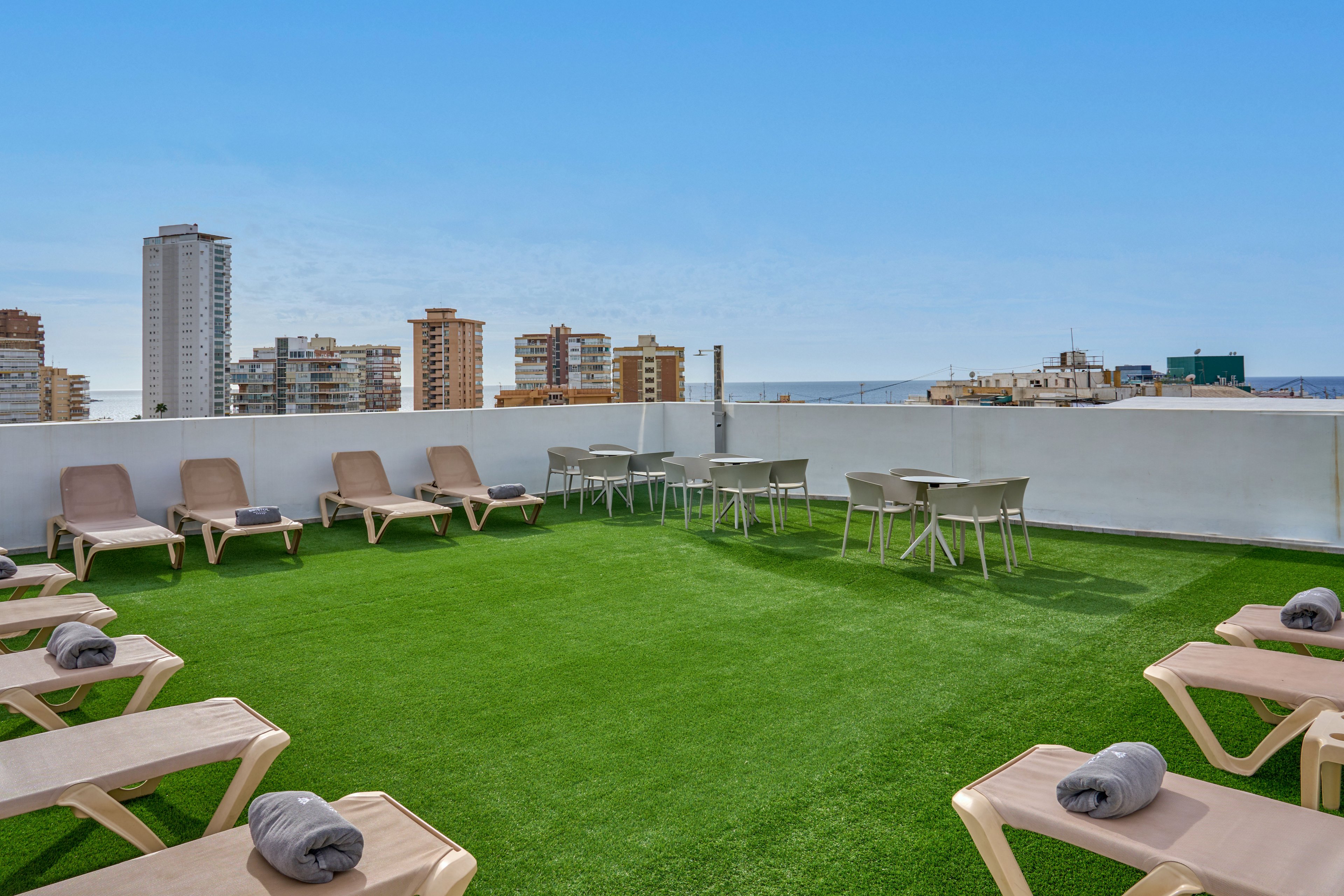 A rooftop terrace with artificial grass features multiple beige sun loungers arranged around the edges. Rolled gray towels rest on each lounger, suggesting a relaxing sunbathing area. In the center, several tables and chairs provide seating with views of surrounding city buildings. The distant sea and clear blue sky create a bright, open atmosphere ideal for leisure.