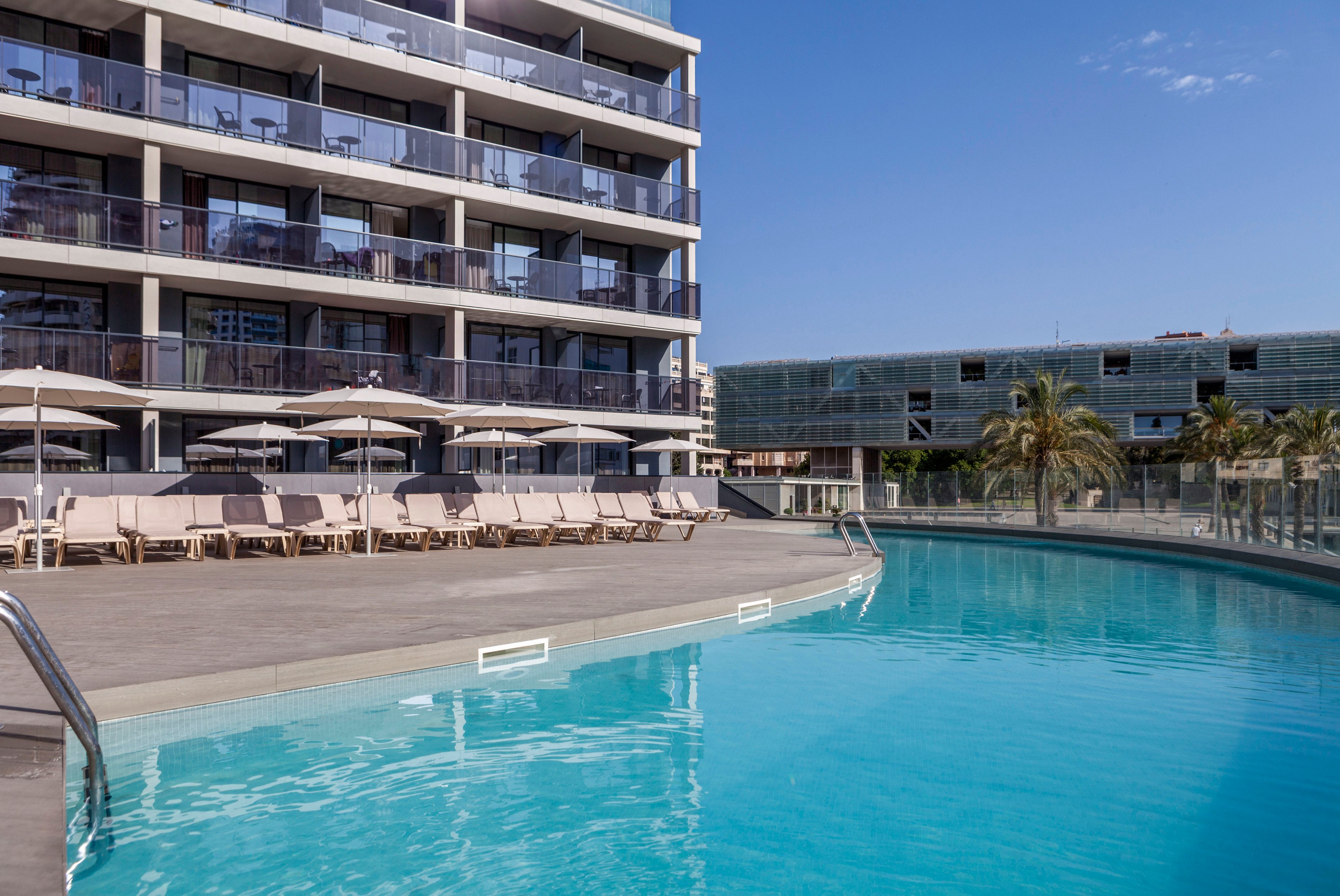 A contemporary multi-story hotel overlooks a clear blue outdoor swimming pool. Sun loungers and umbrellas line the poolside deck, suggesting a relaxing vacation setting. Another building and a few palm trees are visible in the background, adding to the urban resort atmosphere. The scene is captured on a bright, sunny day with a clear sky.