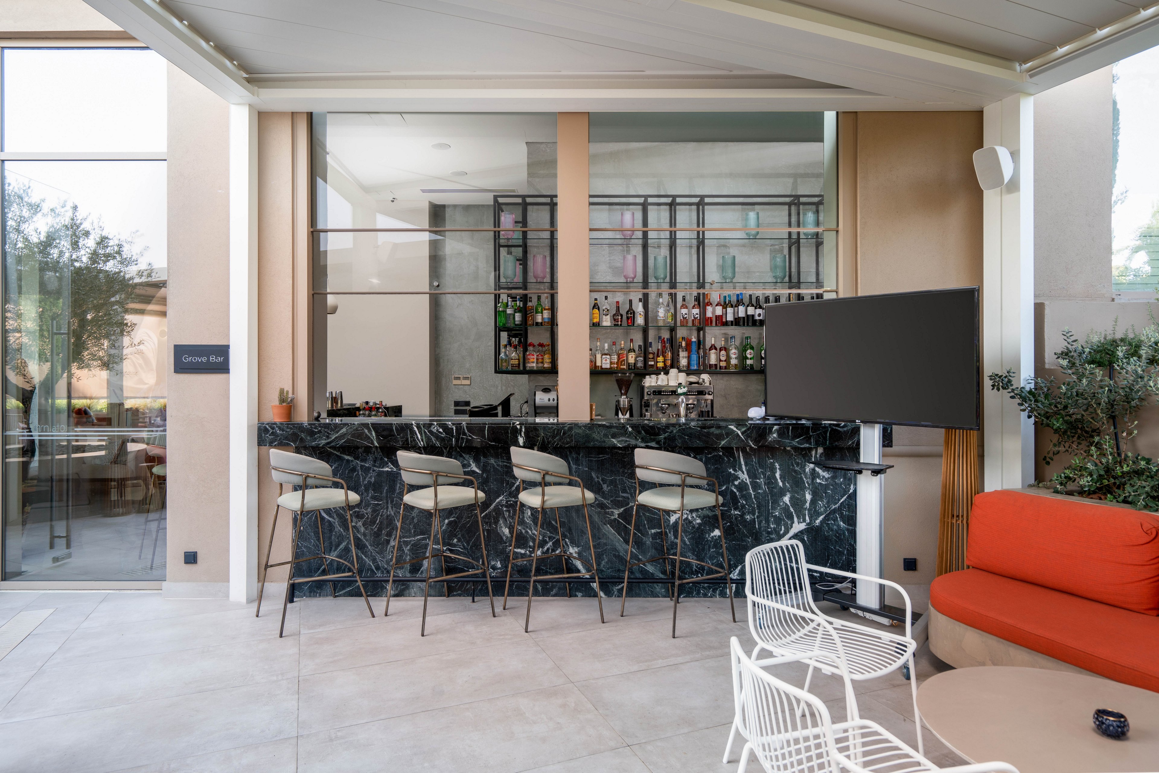 A contemporary outdoor bar features a sleek black marble counter with several high stools neatly arranged along the front. Behind the bar, shelves are stocked with assorted bottles and glassware, creating a refined hospitality setting. The space includes minimalist white wire chairs, a small table, and an orange cushioned bench in the foreground. Large windows, neutral walls, and greenery outside contribute to an airy, upscale atmosphere.