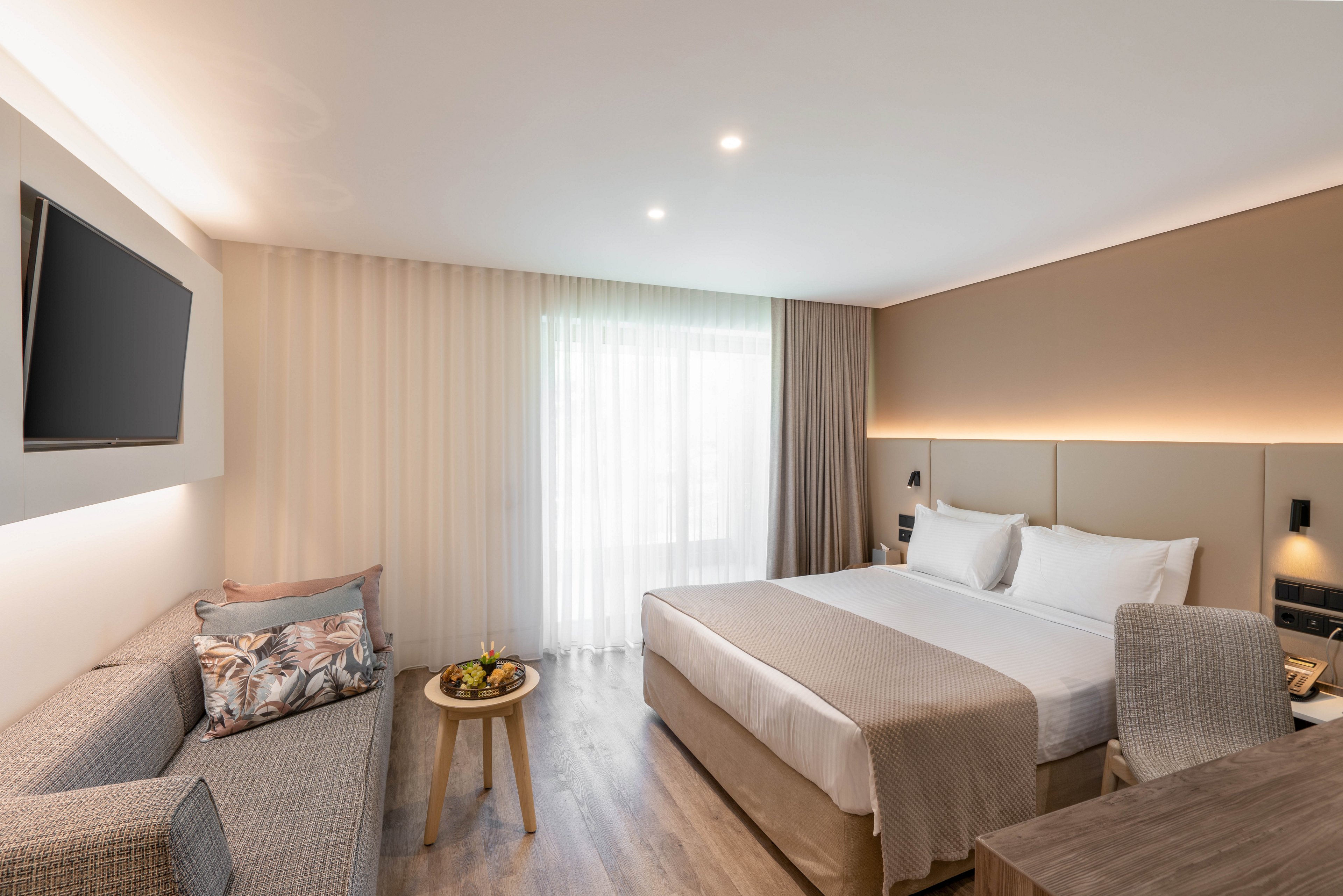 Contemporary hotel room interior featuring a neatly made double bed with neutral bedding. The space includes a gray sofa with patterned cushions, a small round wooden table with a fruit plate, and a wall-mounted flat-screen TV. Soft natural light filters through sheer curtains covering a large window, complemented by warm recessed lighting along the headboard wall. A desk and chair in the foreground suggest a comfortable area for work or study.