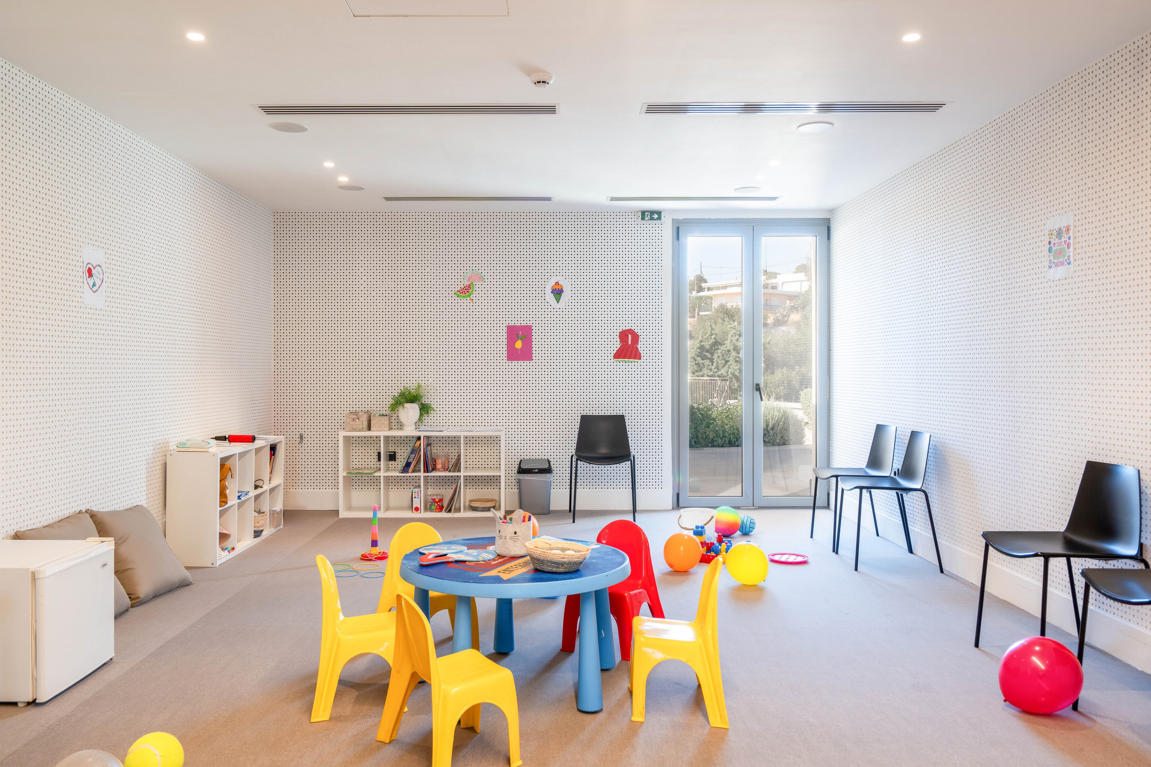 A spacious, modern playroom features a small round blue table surrounded by yellow plastic chairs. Colorful balls and toys are scattered across the light carpeted floor, adding a playful touch. Low shelves with books and storage bins line one wall, while a few black chairs sit neatly against another. Large glass doors at the back let in natural light and offer a view of greenery outside, creating an inviting environment for children.