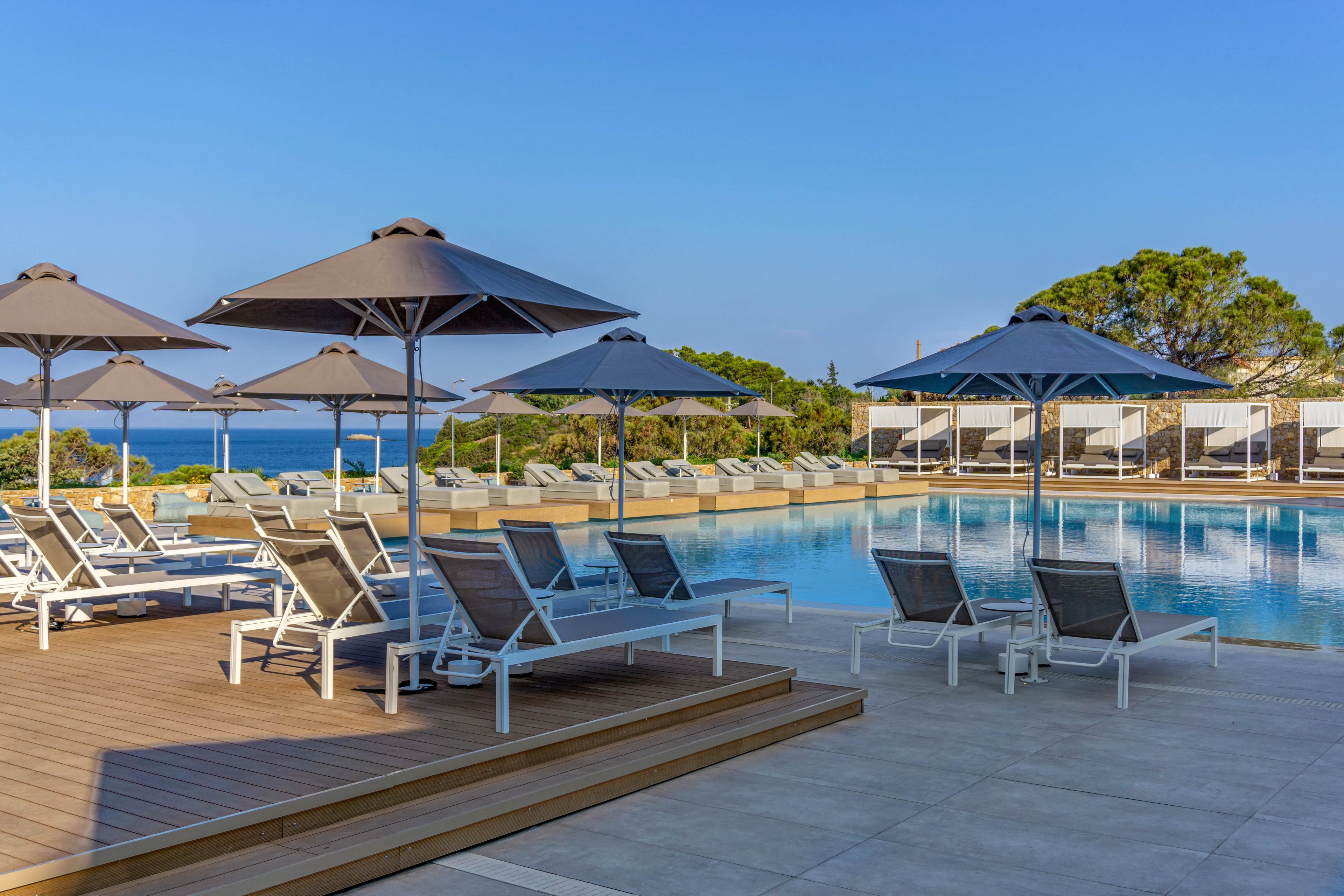 A modern resort swimming pool is surrounded by white sun loungers and large gray umbrellas. The scene is calm and empty, suggesting an early morning or off-peak time at the pool deck. Clear blue water reflects the sky, with the sea and distant trees visible in the background. The overall atmosphere is serene, sunny, and inviting for relaxation.