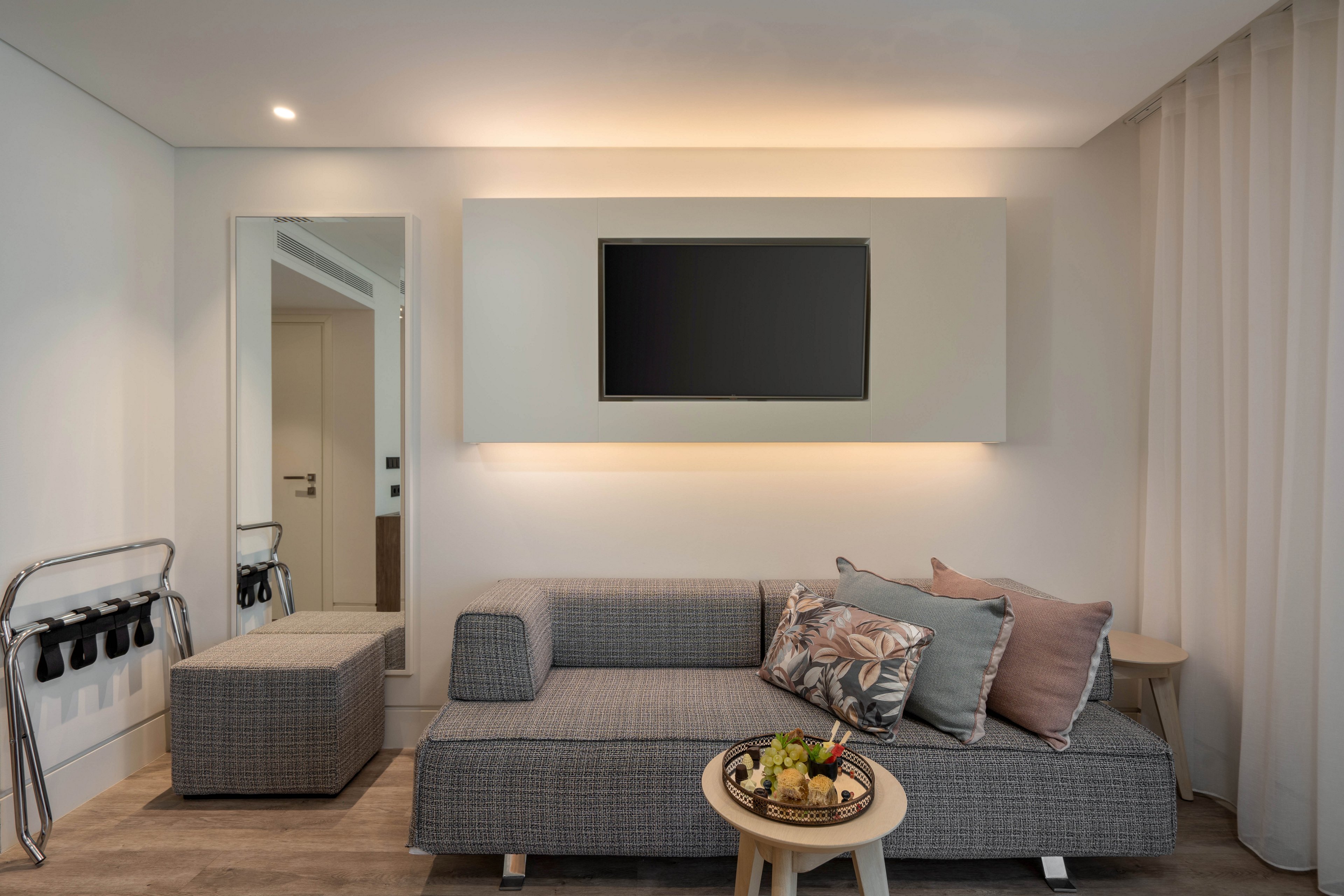 Contemporary living room interior featuring a low gray sofa with patterned and solid throw pillows. A wall-mounted flat screen television is centered above the sofa, framed by soft recessed lighting. A small round side table with a decorative tray and plants sits in the foreground. The space includes a tall mirror, an upholstered ottoman, and light curtains, creating a clean and minimalist atmosphere.