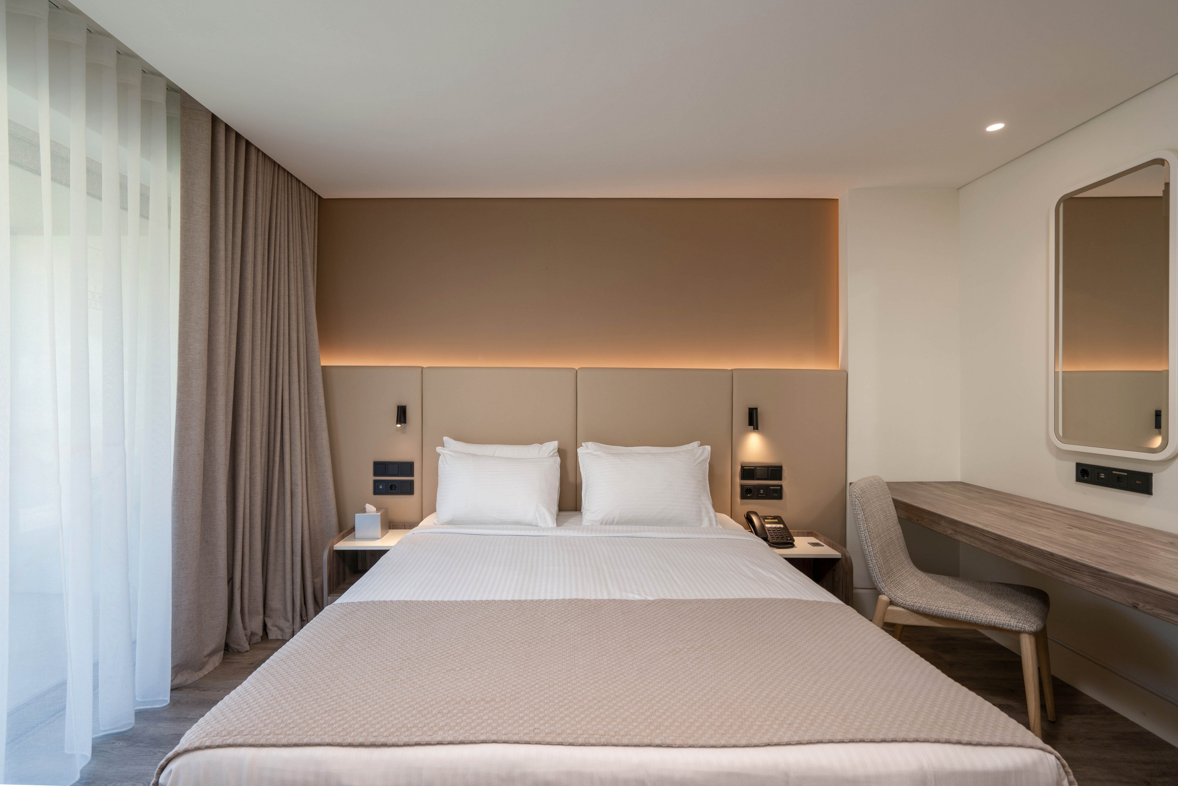 A neatly made double bed with white linens and beige accents stands centered in a modern hotel room. Soft indirect lighting highlights the padded headboard and warm taupe wall behind the bed. Sheer curtains allow natural light to filter in from a large window on the left. A simple desk with a chair, wall mirror, bedside tables, and built-in reading lights complete the minimalist, contemporary design.
