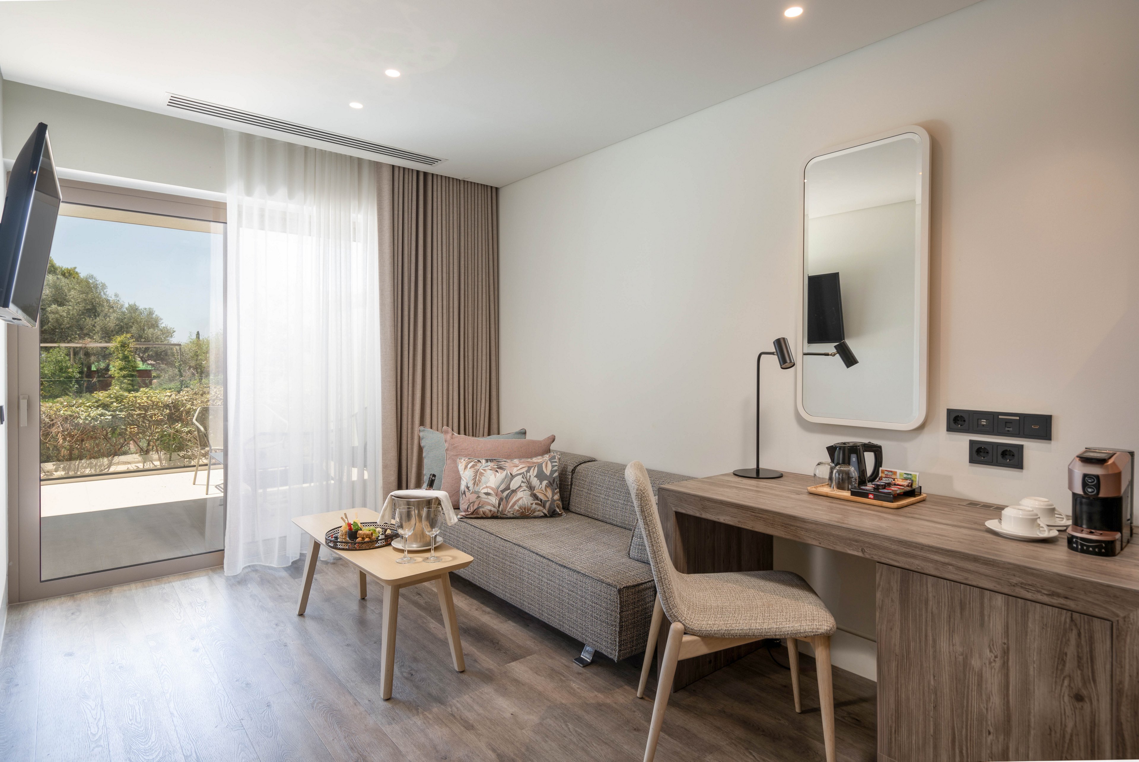 Contemporary hotel room interior featuring a compact gray sofa and wooden coffee table with a breakfast tray. A long wooden desk with a chair, mirror, and desk lamp lines the opposite wall, alongside a coffee maker and cups. Natural light enters through a large glass door with sheer curtains, opening to a terrace and greenery outside. The space is decorated in neutral tones with clean lines, creating a calm and minimalist atmosphere.