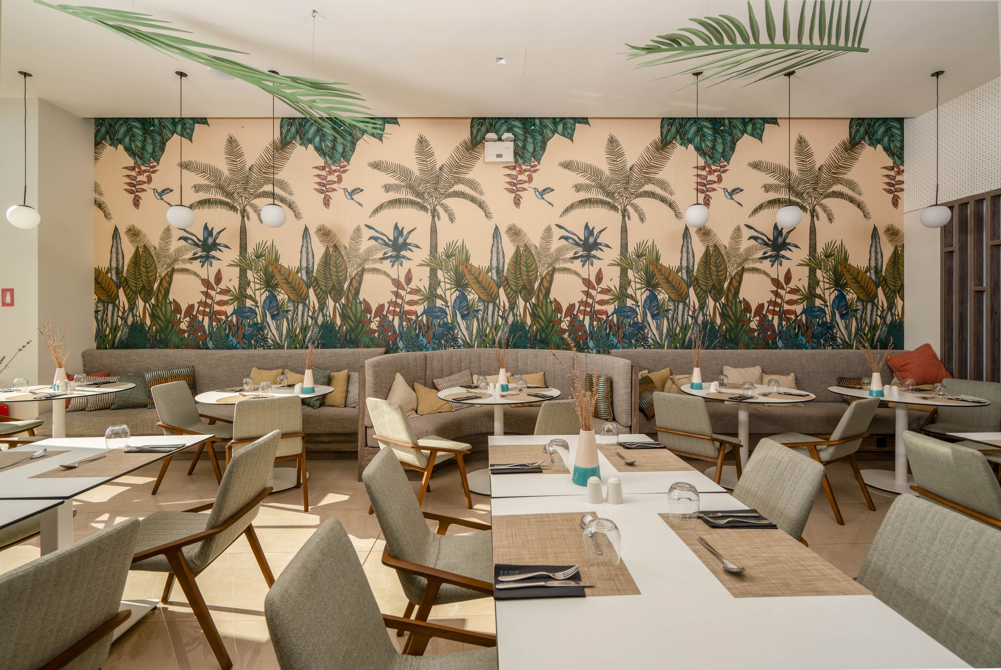 Contemporary restaurant dining room featuring a large tropical mural with palm trees and lush foliage. Light-colored tables and cushioned chairs are neatly arranged, set with glasses, napkins, and simple tableware. Soft neutral tones are accented by green plants and warm wood finishes, creating a relaxed, inviting atmosphere. Pendant lights and natural daylight enhance the airy, modern design of the space.