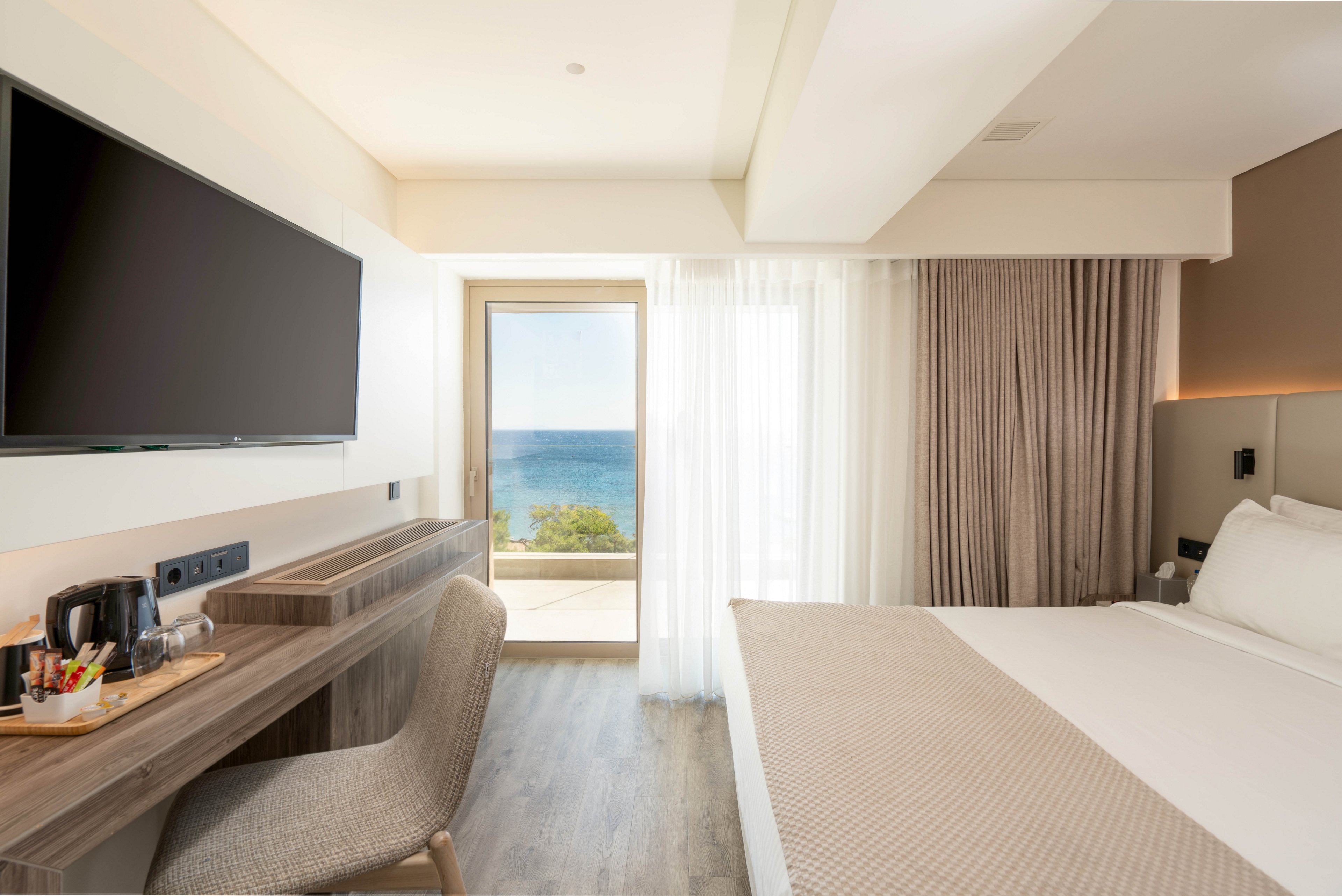 A contemporary hotel room features a neatly made double bed and a wall-mounted flat-screen television. A wooden desk with a chair, electric kettle, and refreshments sits along one side of the room. Floor-to-ceiling curtains frame a glass door that opens to a balcony overlooking the sea. Natural light fills the bright, minimalist interior, creating a calm and inviting atmosphere.