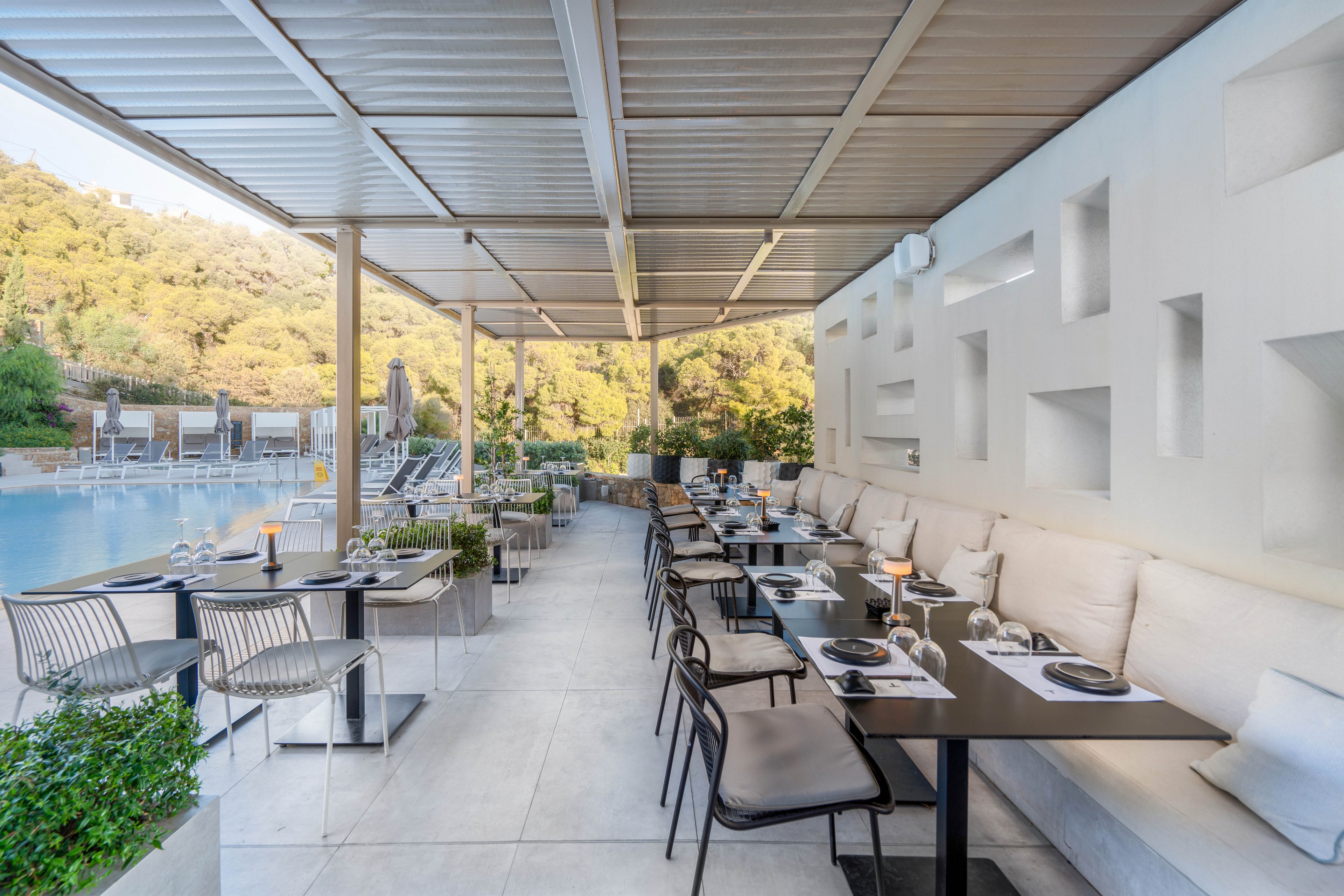 Contemporary outdoor dining tables are arranged neatly along a covered terrace beside a swimming pool. Sleek black tables, metal chairs, and cushioned bench seating create a stylish and comfortable restaurant setting. The space is bright and airy with neutral tones, geometric wall niches, and potted greenery. Hillside trees and pool loungers in the background suggest a resort or hotel environment ideal for relaxed meals.