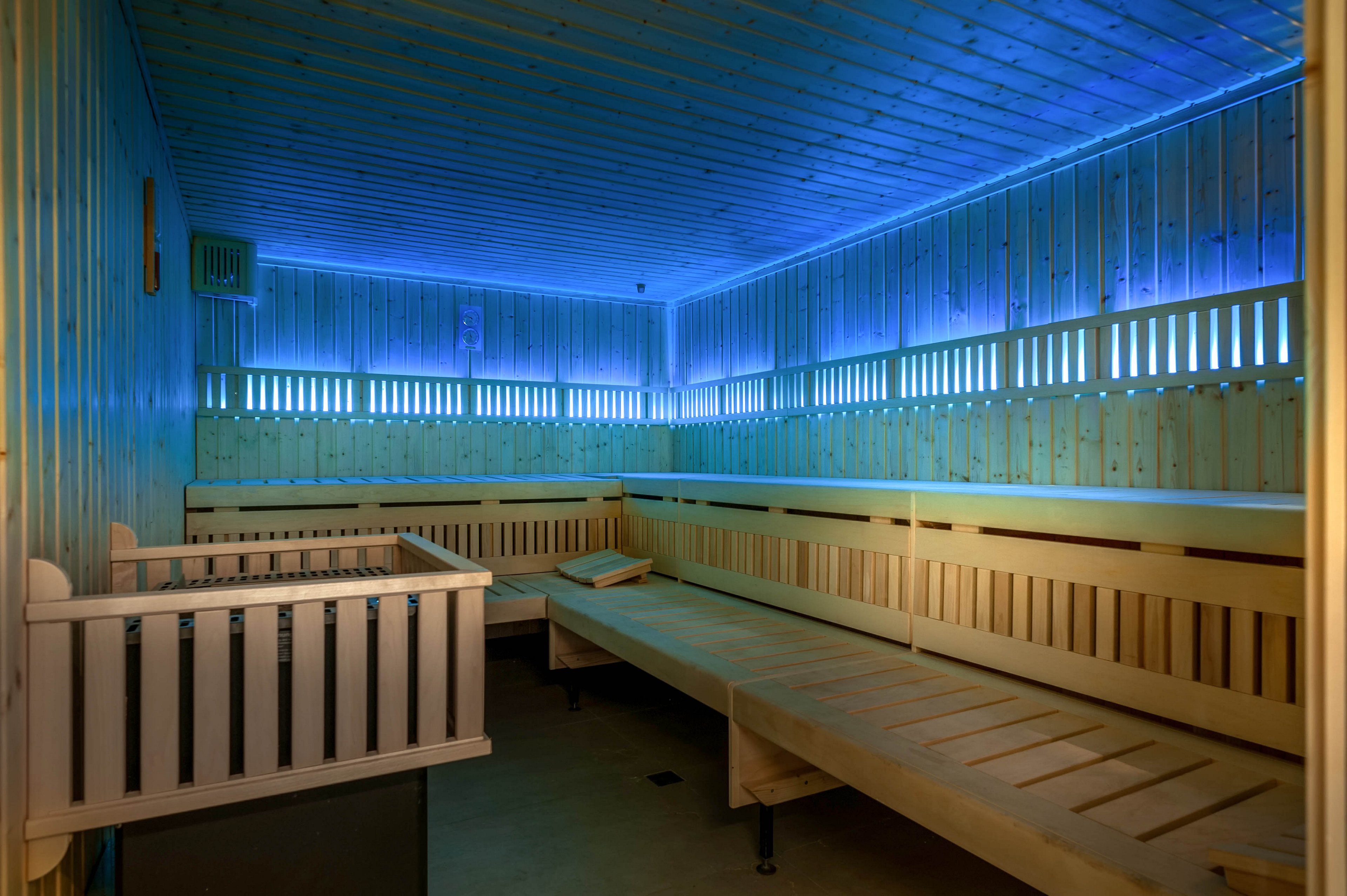 A spacious wooden sauna interior is shown completely empty. Long tiered benches line the walls, creating ample seating space. Soft blue LED lighting runs along the upper walls and ceiling, giving the room a modern, tranquil atmosphere. The clean wood surfaces and warm structure contrast with the cool colored light for a relaxing spa setting.