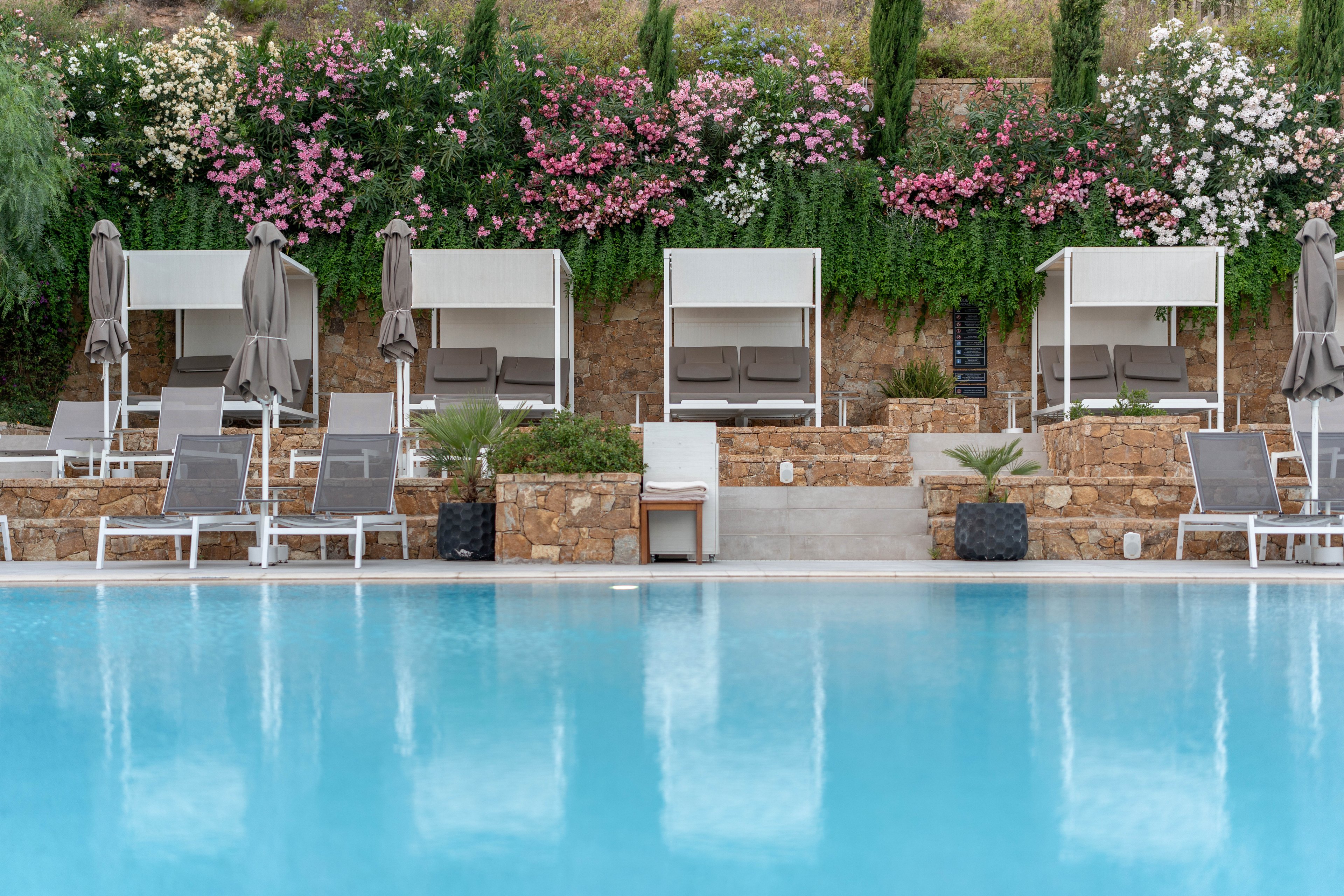A tranquil resort swimming pool is framed by modern white loungers and shaded cabanas. Closed umbrellas line the poolside, suggesting an early morning or off-peak moment of calm. Behind the seating area, stone walls and lush greenery are accented by blooming pink and white flowers. The clear blue water reflects the serene, upscale atmosphere of this outdoor relaxation space.