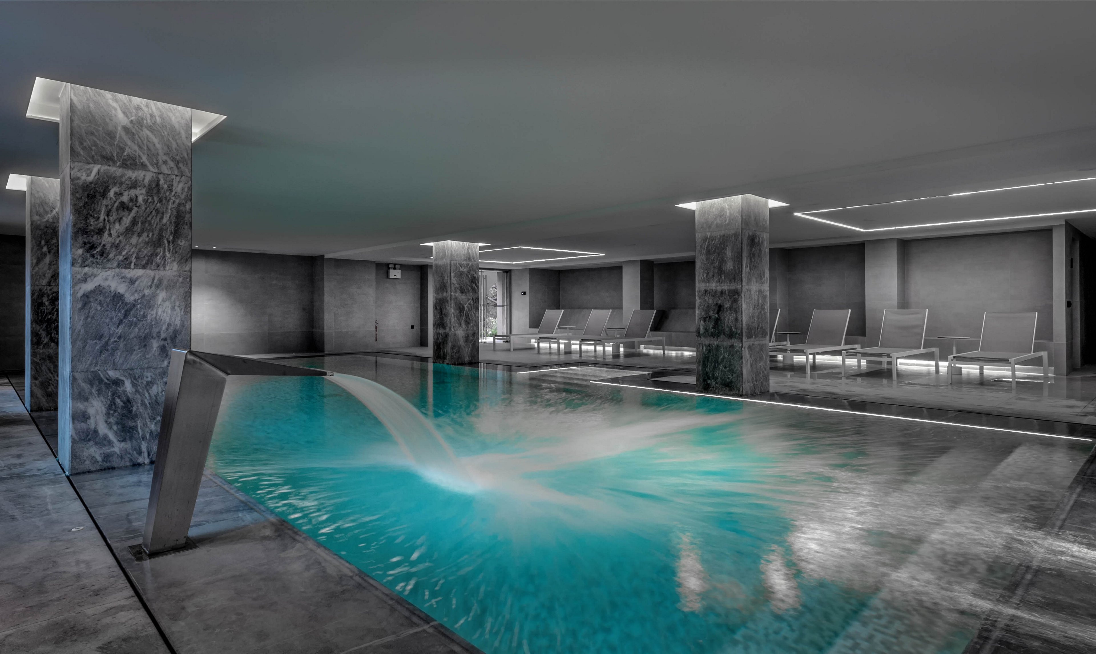 A sleek indoor swimming pool is set within a contemporary spa environment. Water cascades from a stainless steel fountain into the illuminated turquoise pool. Marble-clad columns and grey stone flooring create a minimalist, luxurious atmosphere. In the background, a row of empty lounge chairs lines the wall, emphasizing relaxation and calm. Soft, indirect ceiling lighting enhances the tranquil, high-end design.