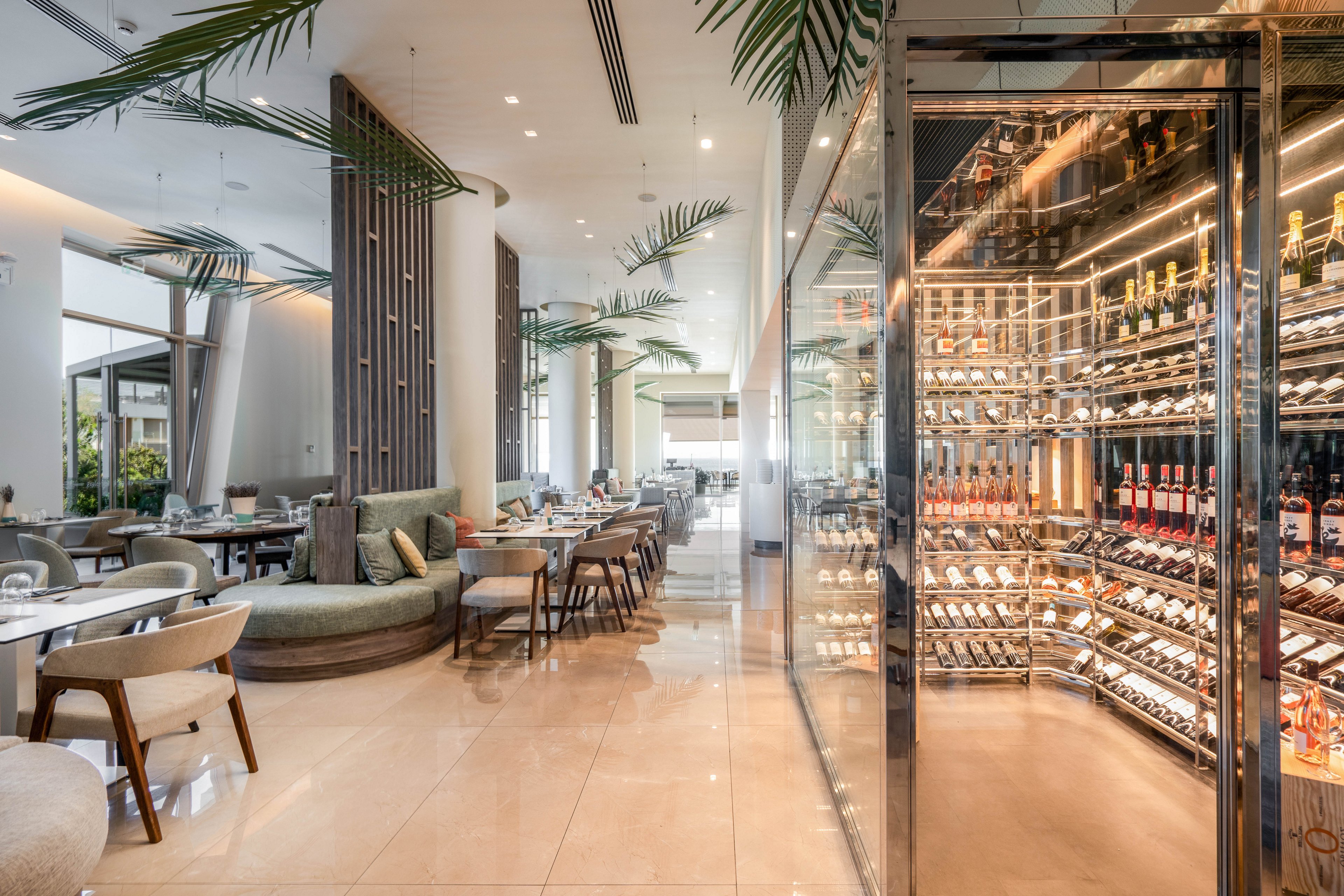 Contemporary restaurant interior features a prominent glass-enclosed wine cellar filled with bottles. The dining area includes round and rectangular tables with upholstered chairs and curved banquette seating. Tall ceilings, large windows, and indoor palm leaves create an airy, upscale atmosphere. Warm lighting reflects off the polished floor, emphasizing the clean, modern design.