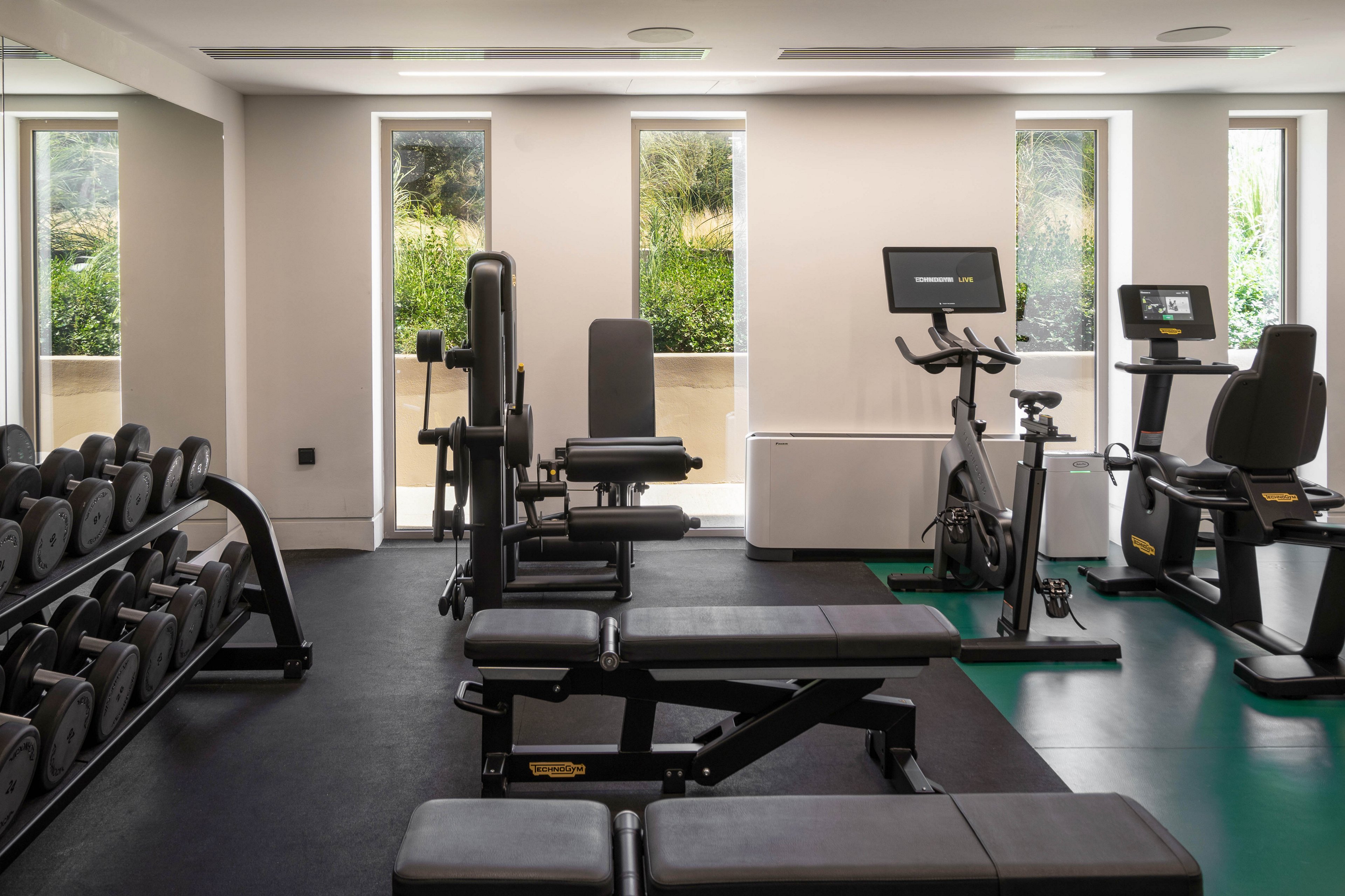A modern indoor gym features various exercise machines and free weights neatly arranged on a dark floor. Benches and strength-training equipment are positioned in the foreground, with dumbbells lined up on racks to the side. Cardio machines with digital screens stand near tall windows that let in natural light and reveal greenery outside. The space appears clean, organized, and designed for individual workouts in a bright, contemporary setting.