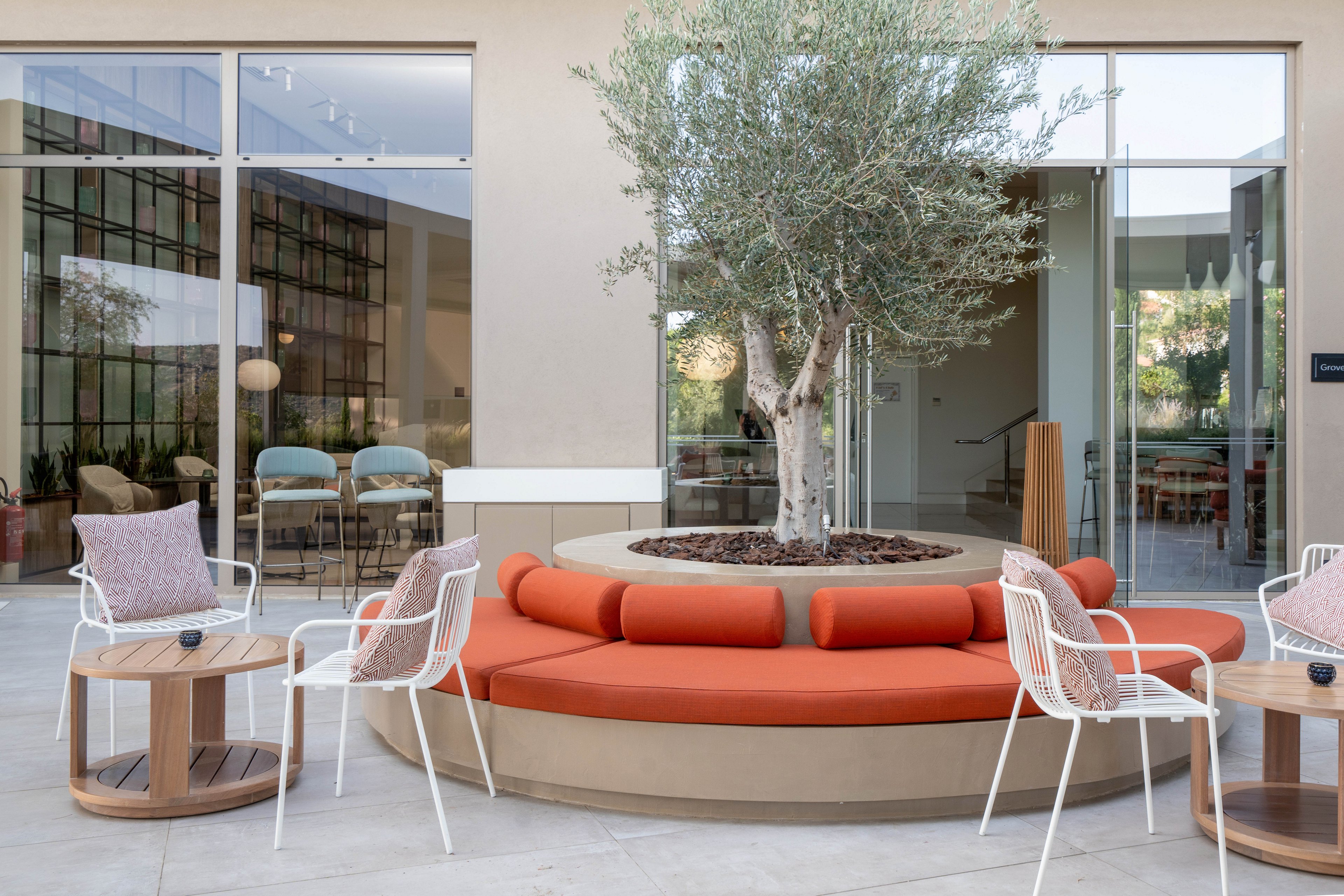 A contemporary outdoor lounge area features a circular bench with orange cushions surrounding a planted tree. White metal chairs and small wooden side tables with cushions and coffee cups create a relaxed seating arrangement. Large glass doors and windows reveal an indoor space with additional seating and modern decor. The neutral architecture and soft color palette emphasize a calm, sophisticated atmosphere.