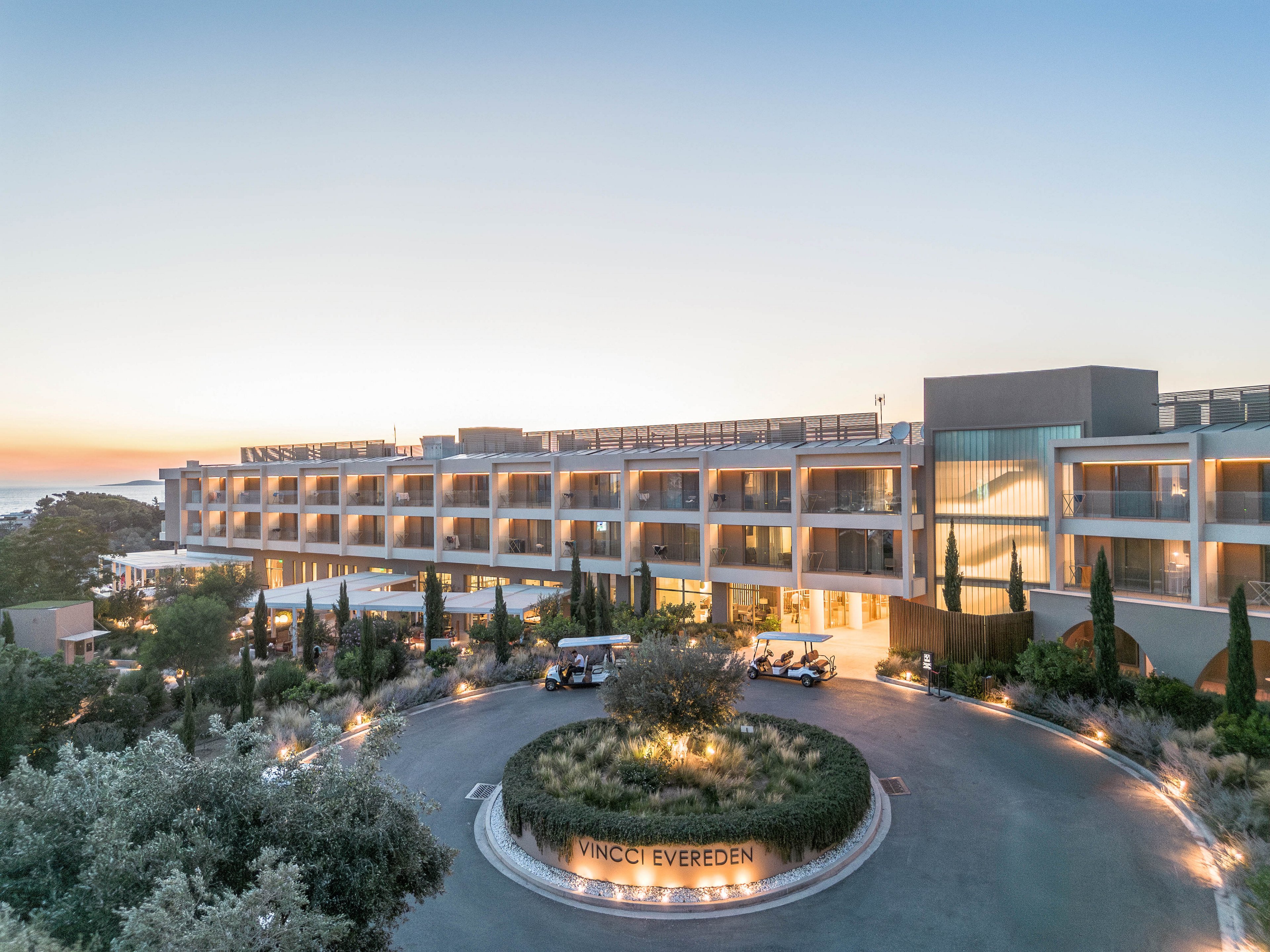 A contemporary multi-story hotel is shown from an elevated viewpoint at sunset, with warm lights illuminating its balconies and facade. The building is surrounded by landscaped gardens, tall cypress trees, and a circular driveway. Several small shuttle carts are parked near the entrance, emphasizing guest transportation and convenience. The sky is clear, and the distant sea is visible on the left, highlighting the coastal location. The text "VINCCI EVEREDEN" appears on the central roundabout feature at the front of the hotel.