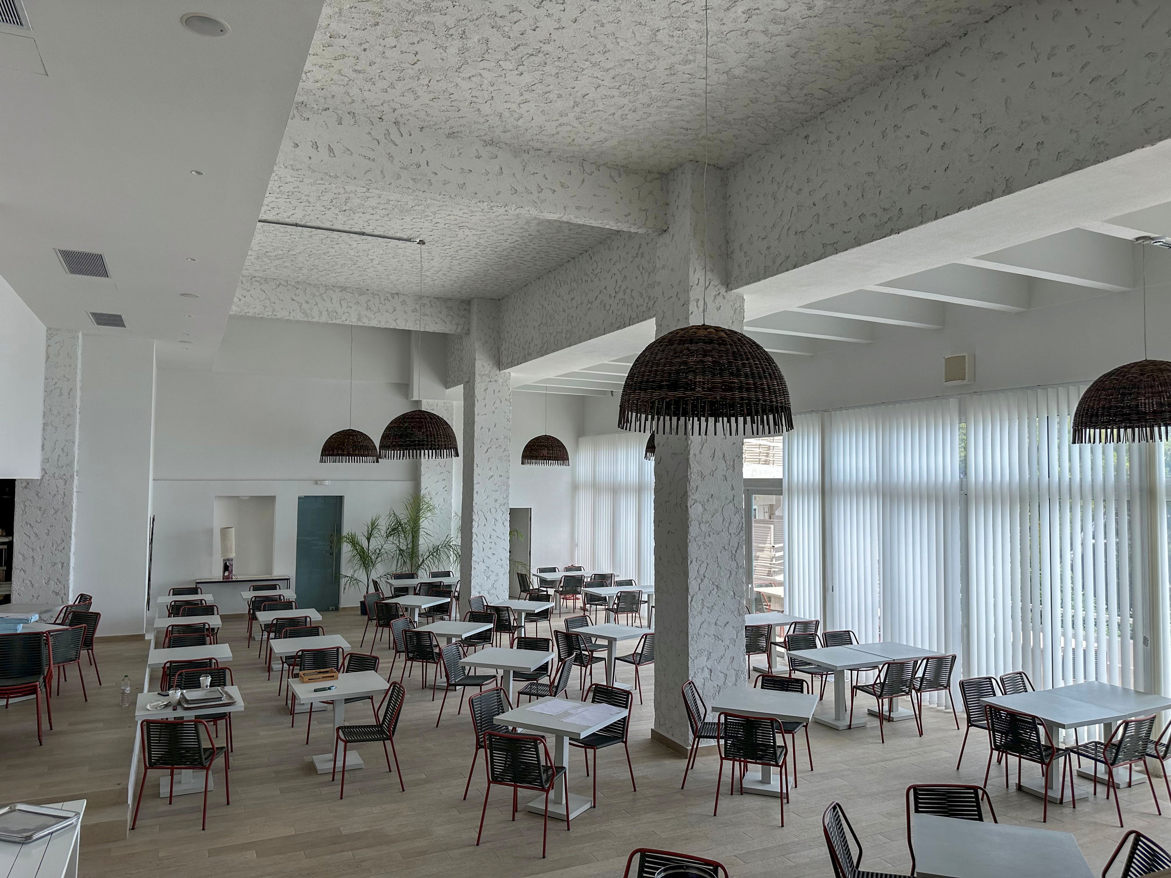 Spacious contemporary restaurant dining area with neatly arranged tables and chairs. Large floor-to-ceiling windows with sheer white curtains allow natural light to fill the room. Woven pendant lights hang from a high patterned ceiling, adding texture and warmth. Light wood flooring and minimal decor create a clean, airy atmosphere suitable for dining or events.