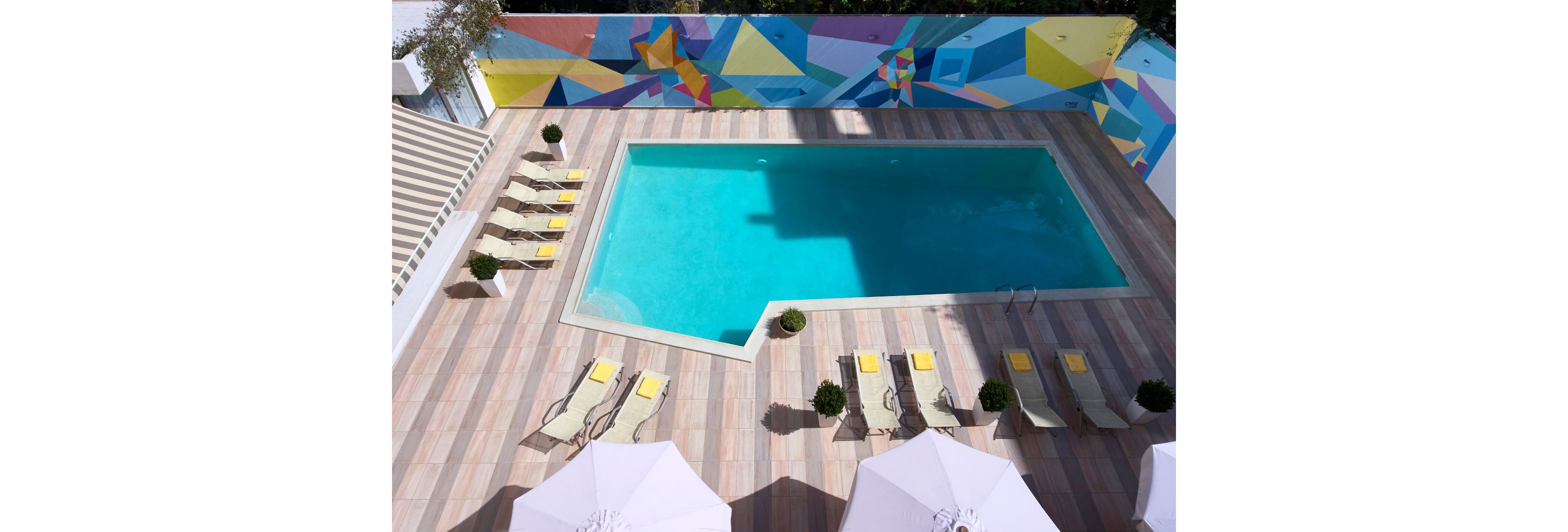 A rectangular outdoor swimming pool surrounded by lounge chairs and umbrellas, set on a tiled deck. The backdrop features a vibrant geometric mural in various colors, adding a lively and artistic touch to the setting. The pool area is well-lit and designed for relaxation and leisure.