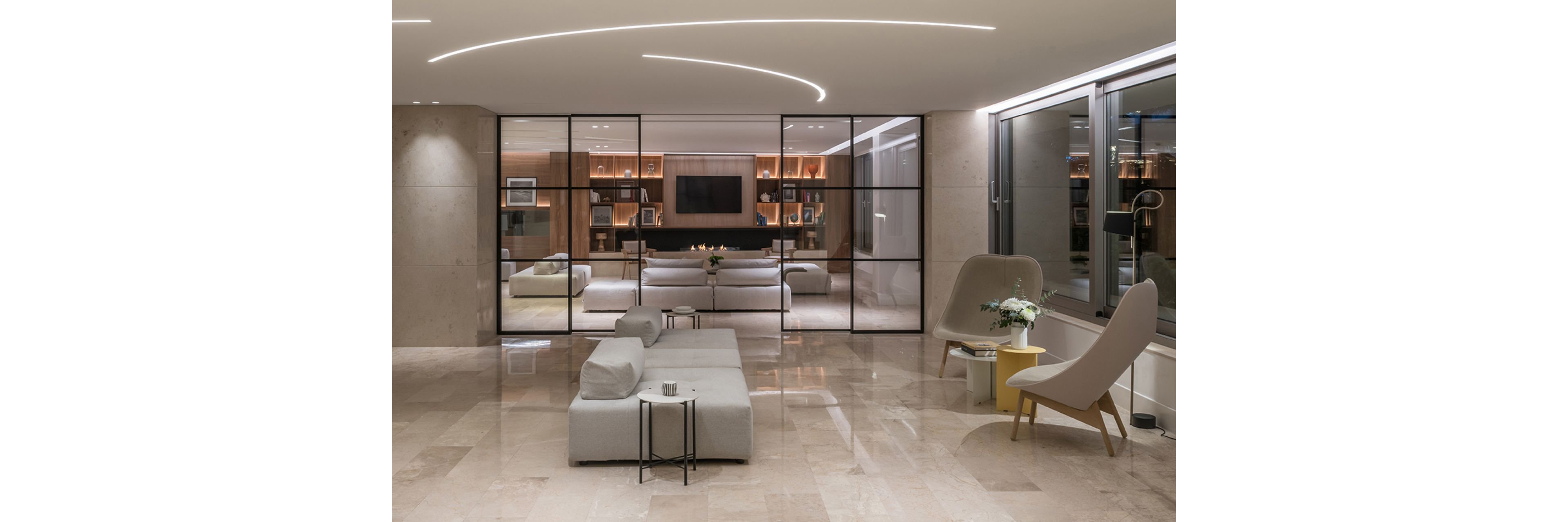 A contemporary lounge featuring minimalist furniture and a spacious layout. The room includes a large glass partition, neutral-toned seating, and decorative accents like flowers and bookshelves. Bright lighting and clean lines enhance the modern aesthetic, creating a sophisticated ambiance.