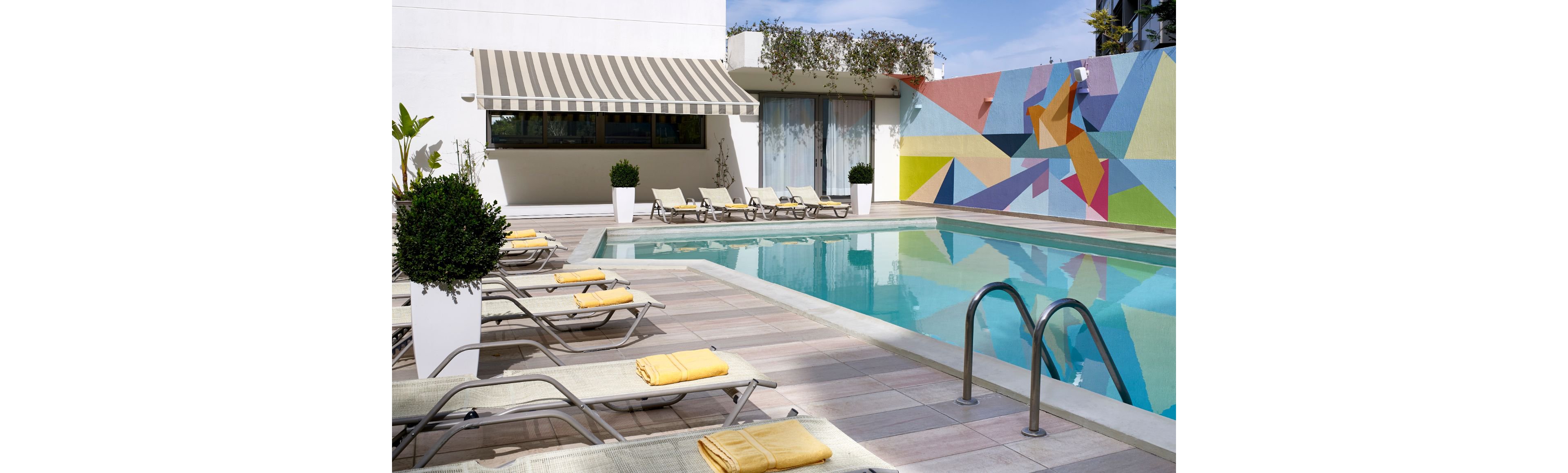 A stylish outdoor pool area featuring lounge chairs with yellow towels, surrounded by a vibrant geometric mural on the wall. The setting includes a striped awning and potted plants, creating a relaxing and contemporary atmosphere. The poolside design emphasizes leisure and modern aesthetics.