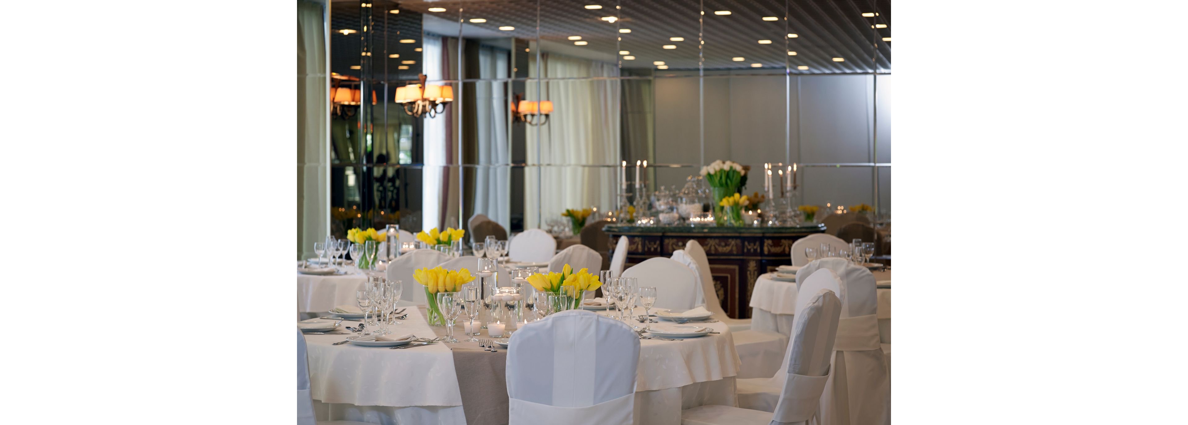 A sophisticated banquet hall featuring round tables adorned with white tablecloths and yellow tulip centerpieces. The mirrored walls and soft lighting create a luxurious ambiance. Glassware, candles, and decorative accents enhance the refined setting, ideal for formal events or celebrations.