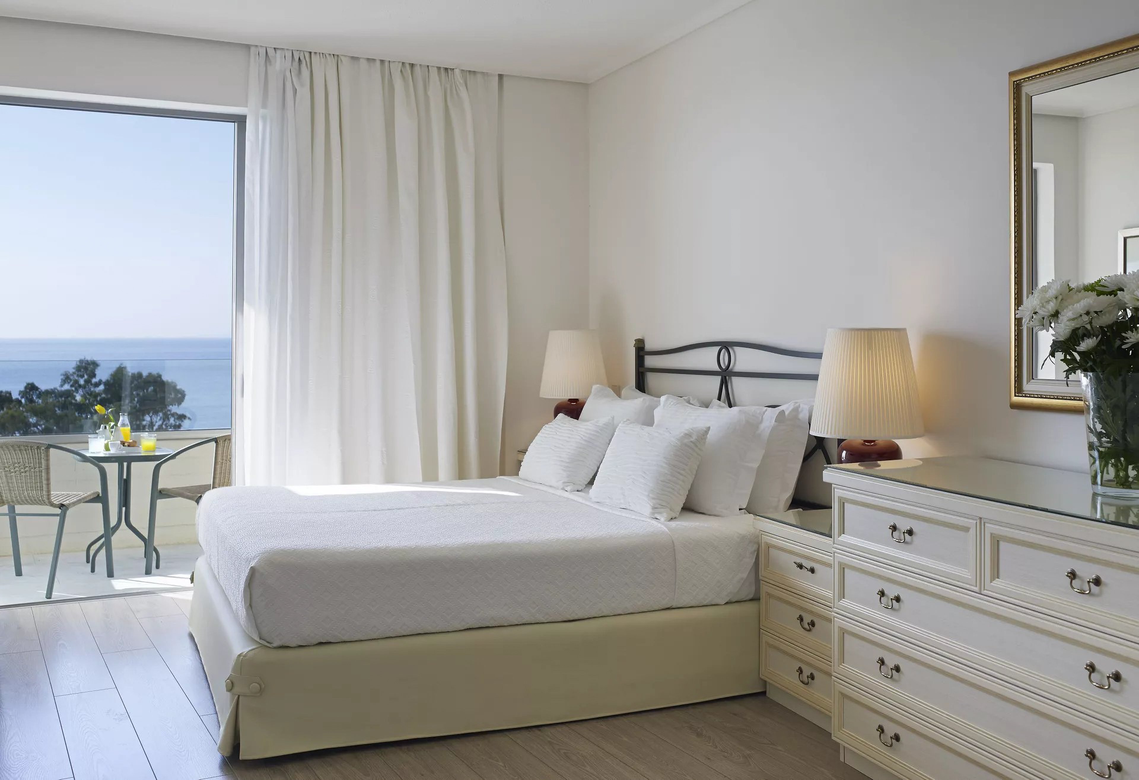 A serene bedroom featuring a neatly made bed with white linens, a dresser, and soft lighting from table lamps. The room opens to a balcony with a seating area overlooking a calm ocean view. The decor is minimalist with a neutral palette, creating a relaxing atmosphere.