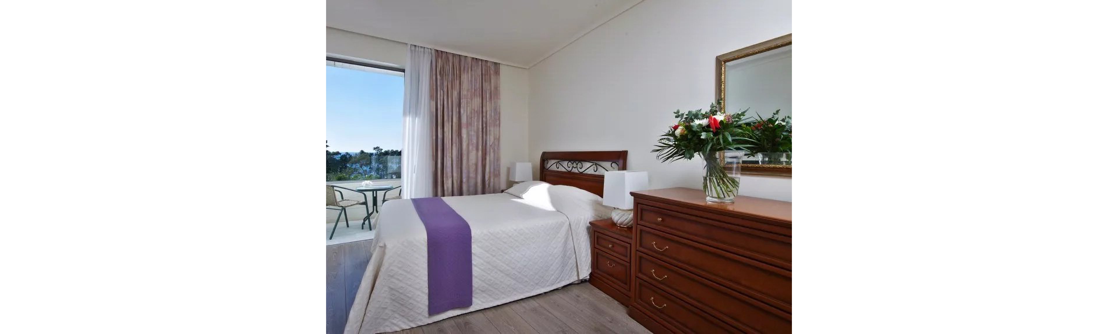 A well-lit bedroom featuring a neatly made bed with a white quilt and purple accent runner. The room includes wooden furniture, such as a dresser and bedside tables, and a vase of fresh flowers. A sliding glass door opens to a balcony with outdoor seating and a scenic view.