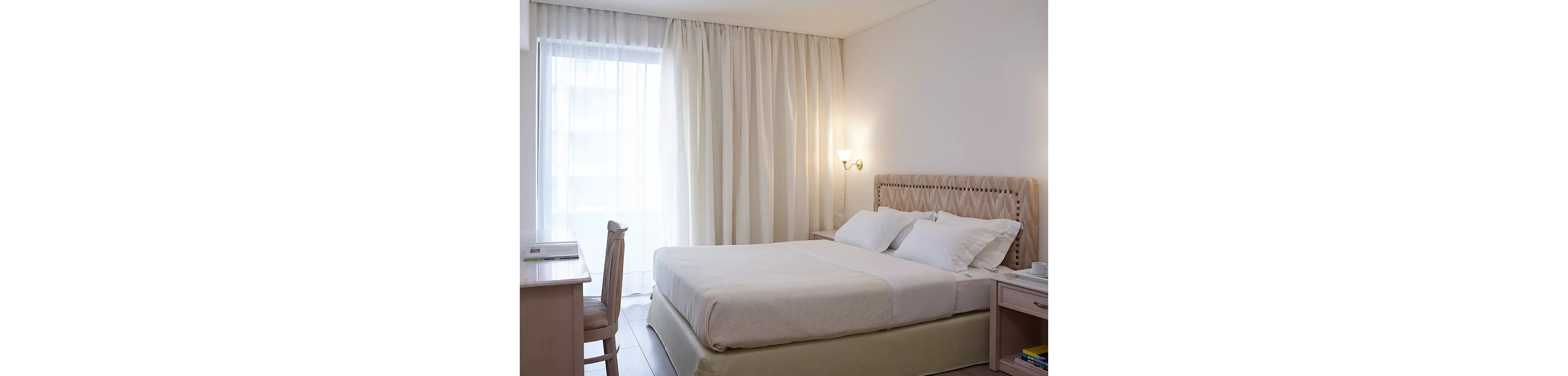 A well-lit hotel room featuring a neatly made double bed with white linens. The room includes a wooden desk and chair near a large window with sheer curtains, allowing natural light to fill the space. Warm wall lighting adds to the cozy and inviting atmosphere.