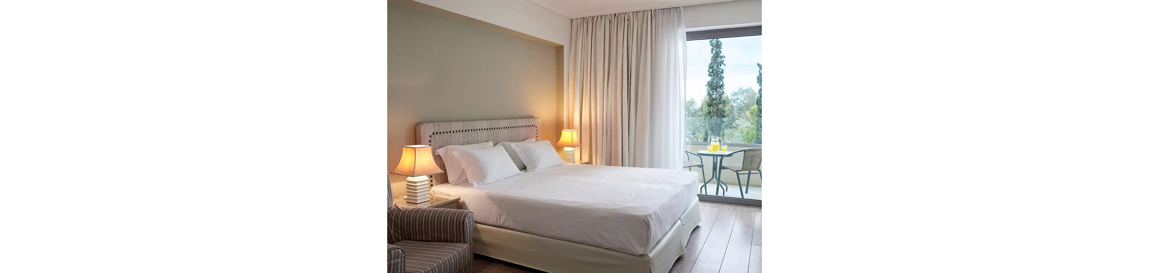 A well-lit hotel room featuring a neatly made double bed with white linens, two bedside lamps, and a striped armchair. The room opens to a balcony with a small table, chairs, and a view of trees. The neutral color palette and soft lighting create a calm and inviting atmosphere.