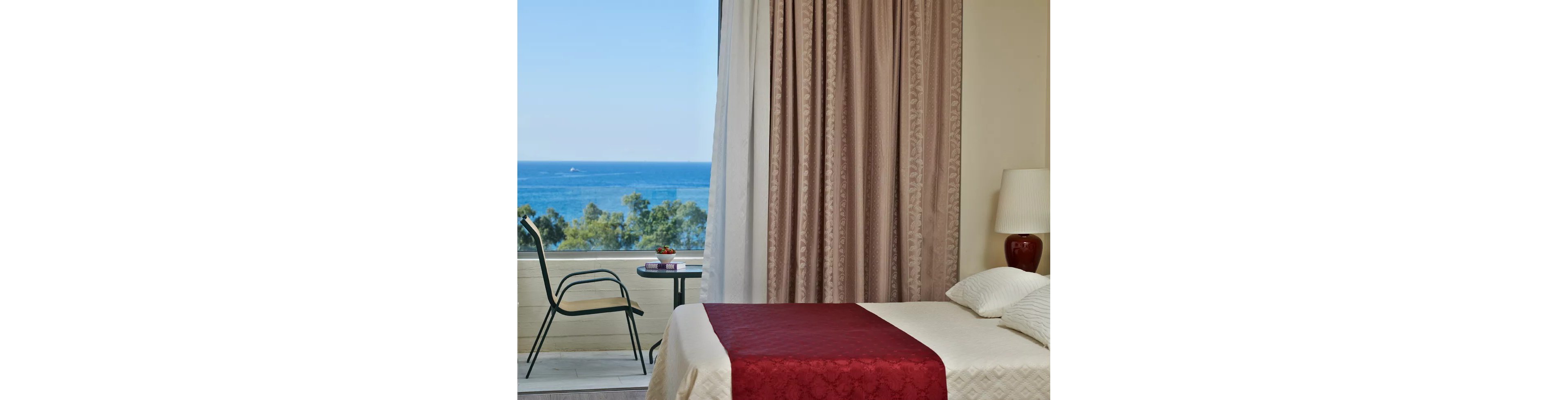 A well-lit hotel room featuring a neatly made bed with a red bedspread and white pillows. The room opens to a balcony with a table and chair, offering a stunning view of the ocean and greenery. Soft curtains frame the large window, adding a touch of elegance to the serene setting.