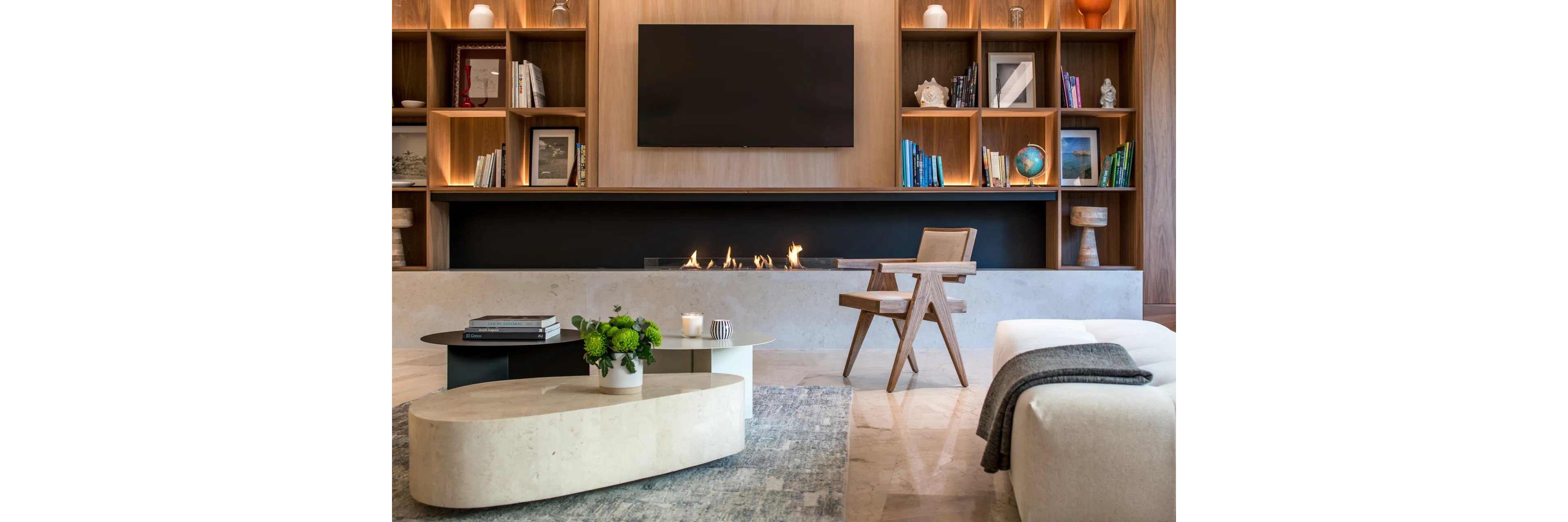 A contemporary living room featuring a sleek wall-mounted fireplace beneath a large flat-screen TV. Wooden shelving units with books and decor items add warmth and character to the space. The room includes a minimalist coffee table, a wooden chair, and a cozy sofa with a throw blanket. Neutral tones and soft lighting create a calm and inviting atmosphere.