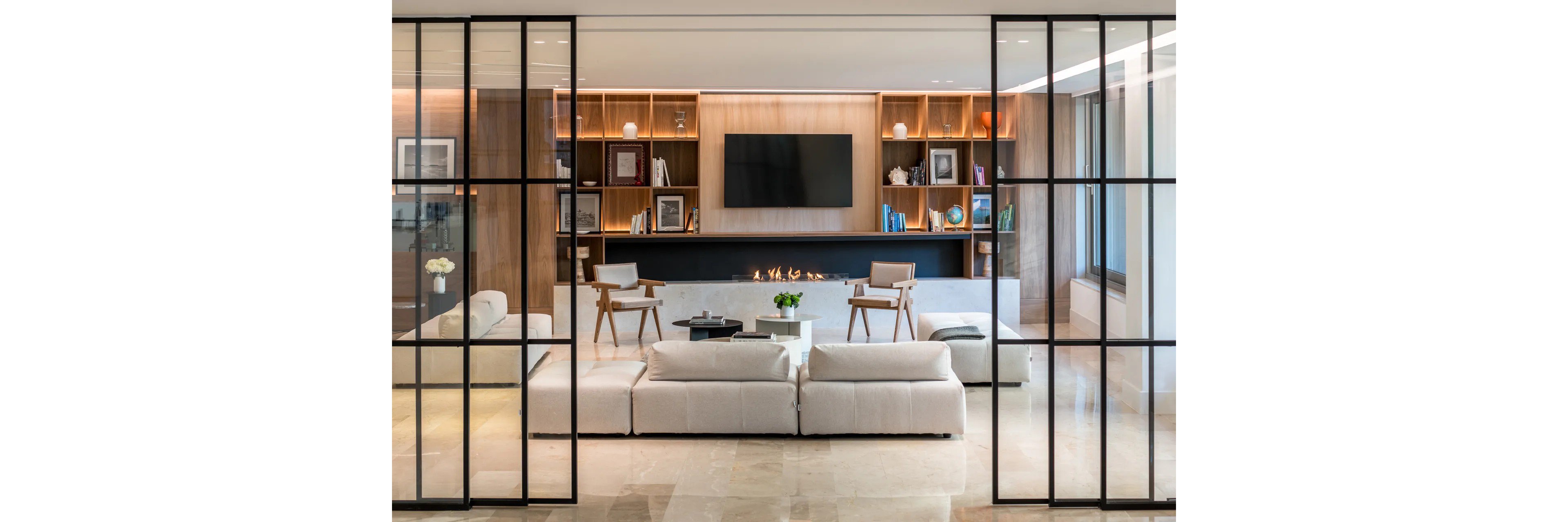 A contemporary living room featuring sleek glass partitions and a cozy seating arrangement. The space includes white sofas, wooden chairs, and a built-in fireplace beneath a wall-mounted TV. Shelves with books and decor items add warmth and character to the minimalist design.