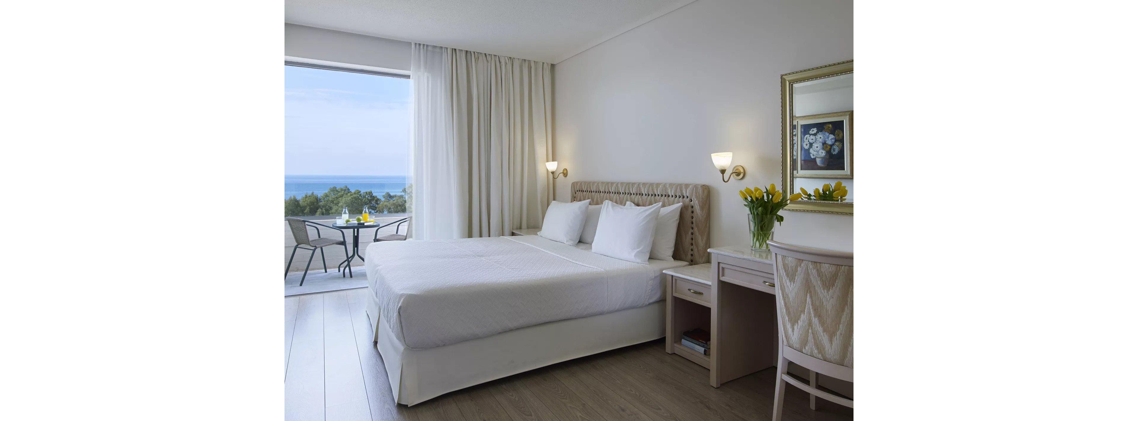 A modern hotel bedroom features a large neatly made bed with white linens and multiple pillows. The room is decorated in light neutral tones with wooden furniture, including a desk, chair, and bedside table. A vase of yellow tulips and framed artwork add subtle decorative accents. Floor-to-ceiling curtains open to a balcony with a small table and chairs overlooking the sea and distant coastline.