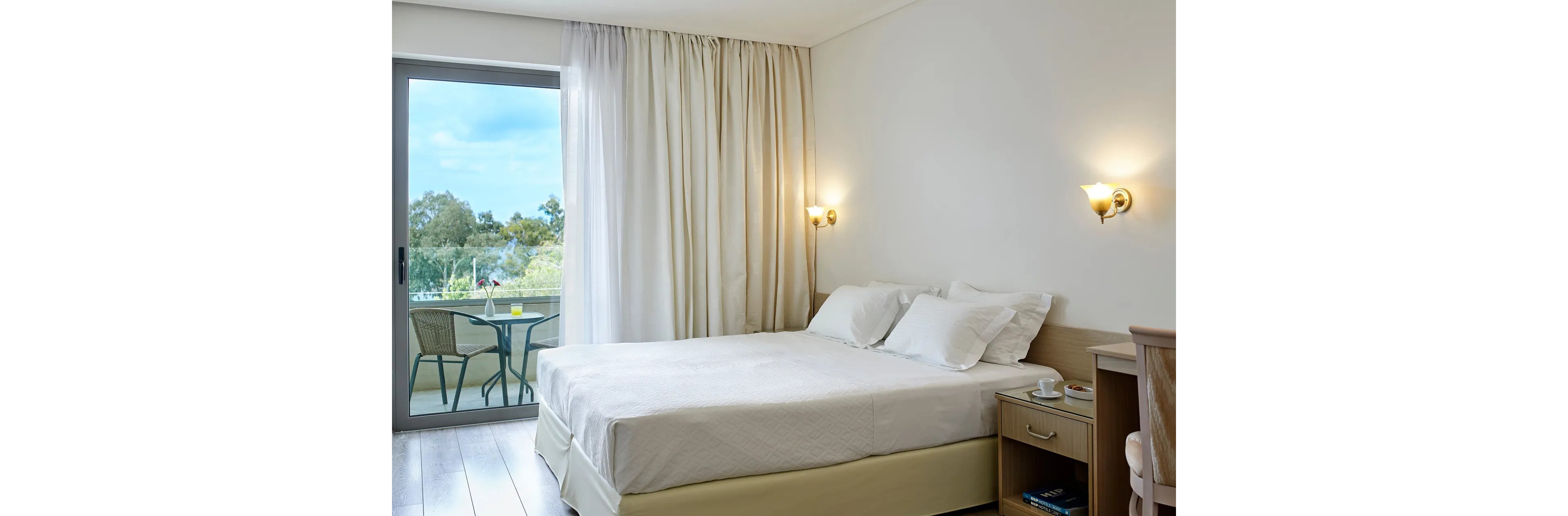 A neatly made double bed with white linens is placed against a light-colored wall in a modern hotel room. Large curtains frame a sliding glass door that opens to a balcony with a table and two chairs overlooking greenery. A wooden nightstand with a lamp, phone, and shelves sits beside the bed, adding functional comfort. Soft natural light and neutral tones create a calm, inviting atmosphere ideal for relaxation.