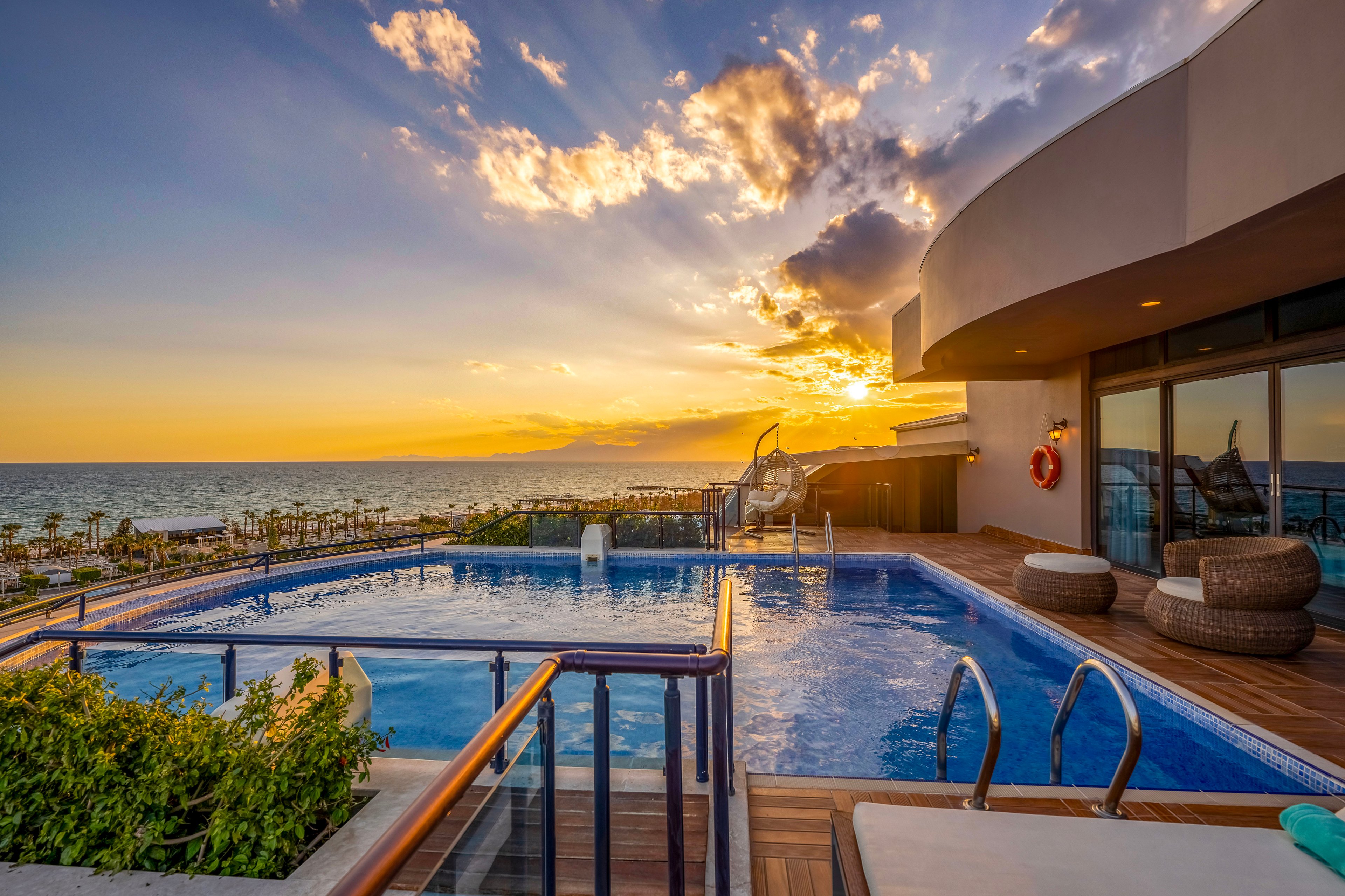A modern rooftop infinity pool overlooks a calm ocean at sunset, with golden light reflecting on the water. Curved contemporary architecture frames the terrace, featuring lounge chairs and round wicker seating. The sky is filled with dramatic clouds glowing in warm orange and blue tones. Palm trees and coastal scenery stretch along the shoreline in the distance, creating a serene vacation atmosphere.