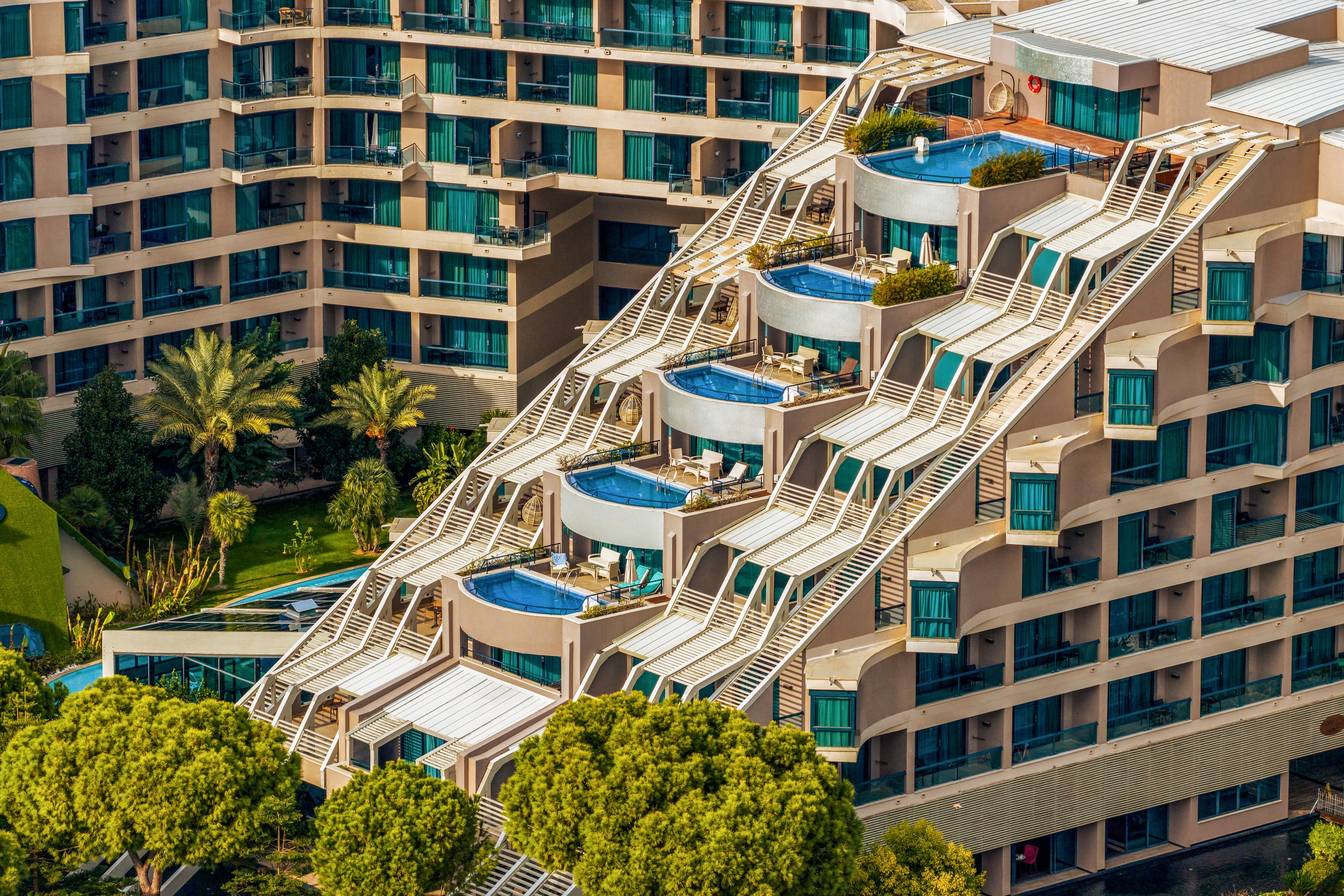 A modern terraced hotel features multiple private rooftop swimming pools cascading down its stepped facade. The building’s contemporary architecture includes curved balconies, white structural beams, and large glass windows. Lush green trees and palm trees surround the property, adding a resort-like atmosphere. The scene is captured from above, highlighting the geometric design and vibrant blue water of the pools.
