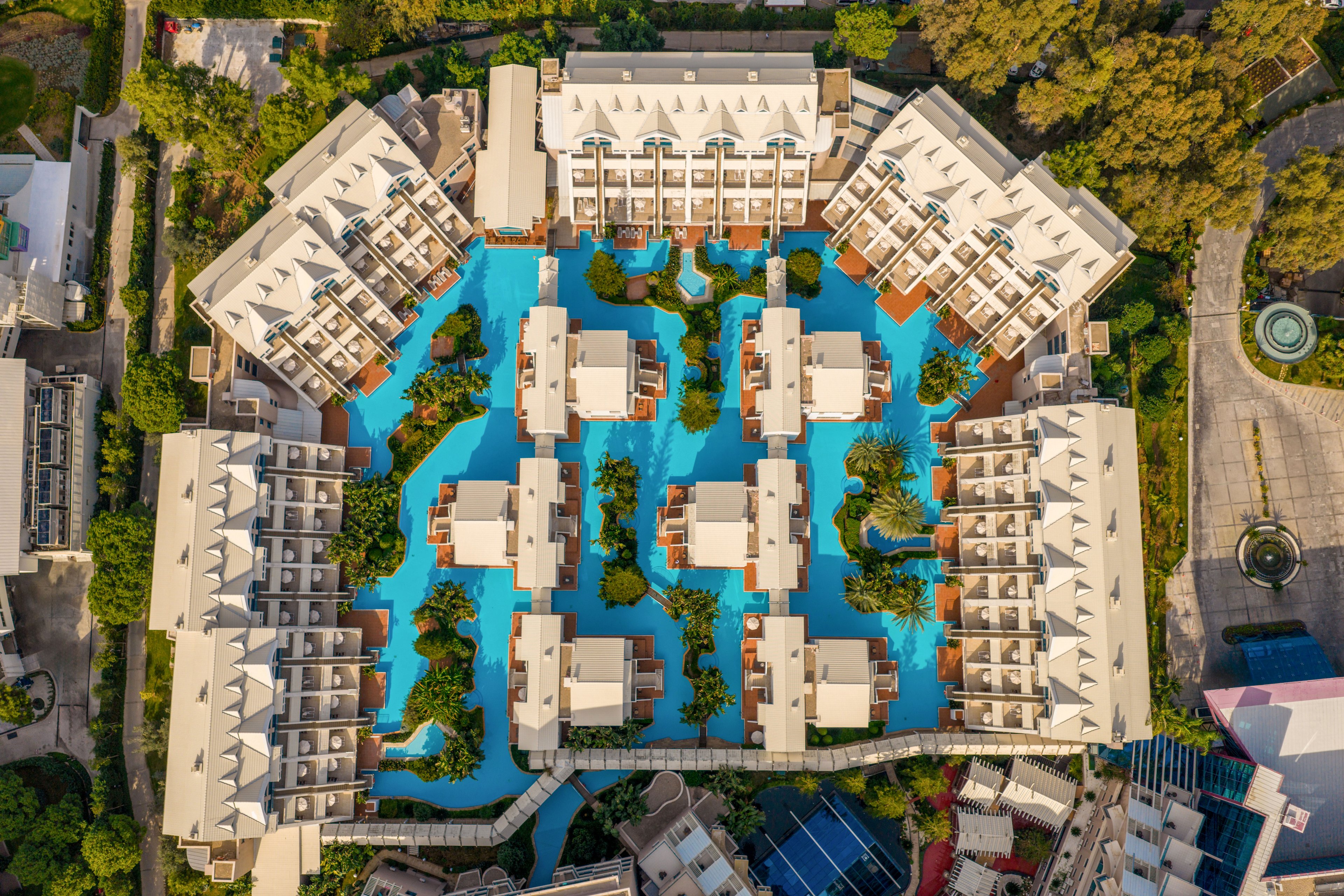 A large resort complex is photographed from above, revealing a symmetrical layout of buildings surrounding an expansive central swimming pool. Multiple white-roofed structures are arranged in geometric patterns with walkways connecting them. The bright blue water contrasts with the neutral architecture and surrounding greenery. Nearby outdoor areas and sports courts are partially visible at the edges of the frame.
