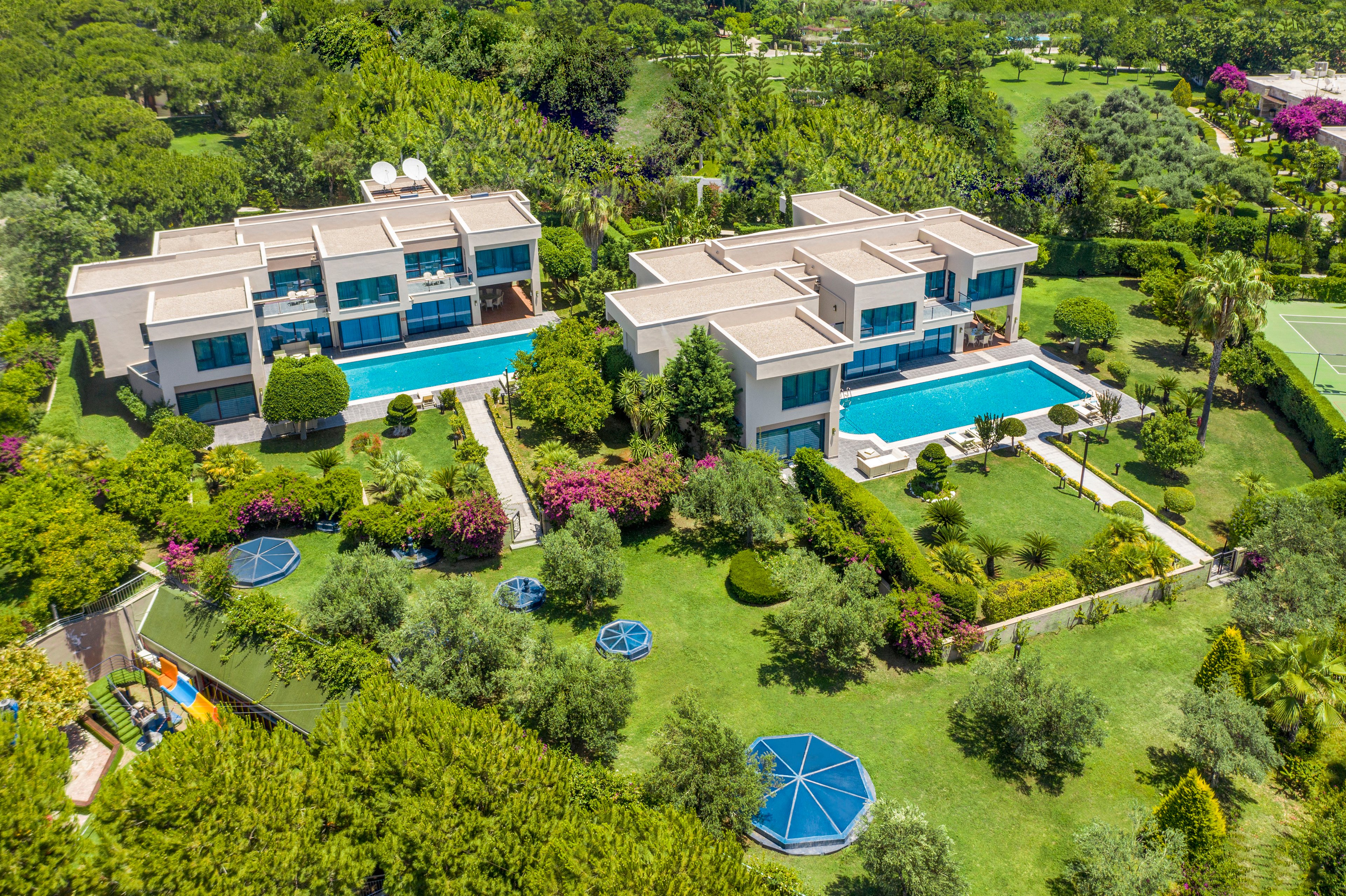 A pair of modern luxury villas with large rectangular swimming pools are seen from above in a lush green landscape. The properties are surrounded by manicured lawns, flowering shrubs, and mature trees, creating a resort-like atmosphere. Blue circular shade structures and garden paths add visual interest to the expansive grounds. In the background, more greenery and distant buildings suggest a secluded residential or vacation area.