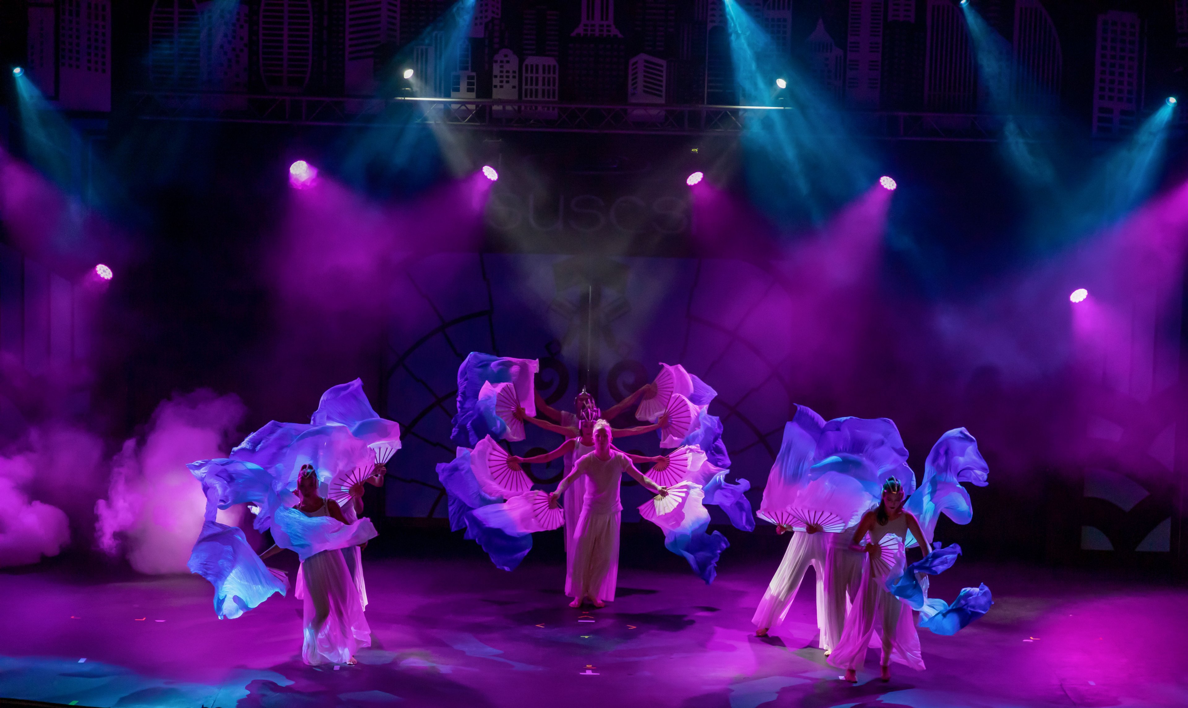 A group of dancers performs on a theatrical stage under dramatic purple and blue lighting. They wear long white costumes and wave large blue translucent fabric pieces that create dynamic shapes around them. Smoke effects and a stylized cityscape backdrop enhance the performance atmosphere. The overall scene feels energetic and artistic, emphasizing movement, color, and coordinated choreography.