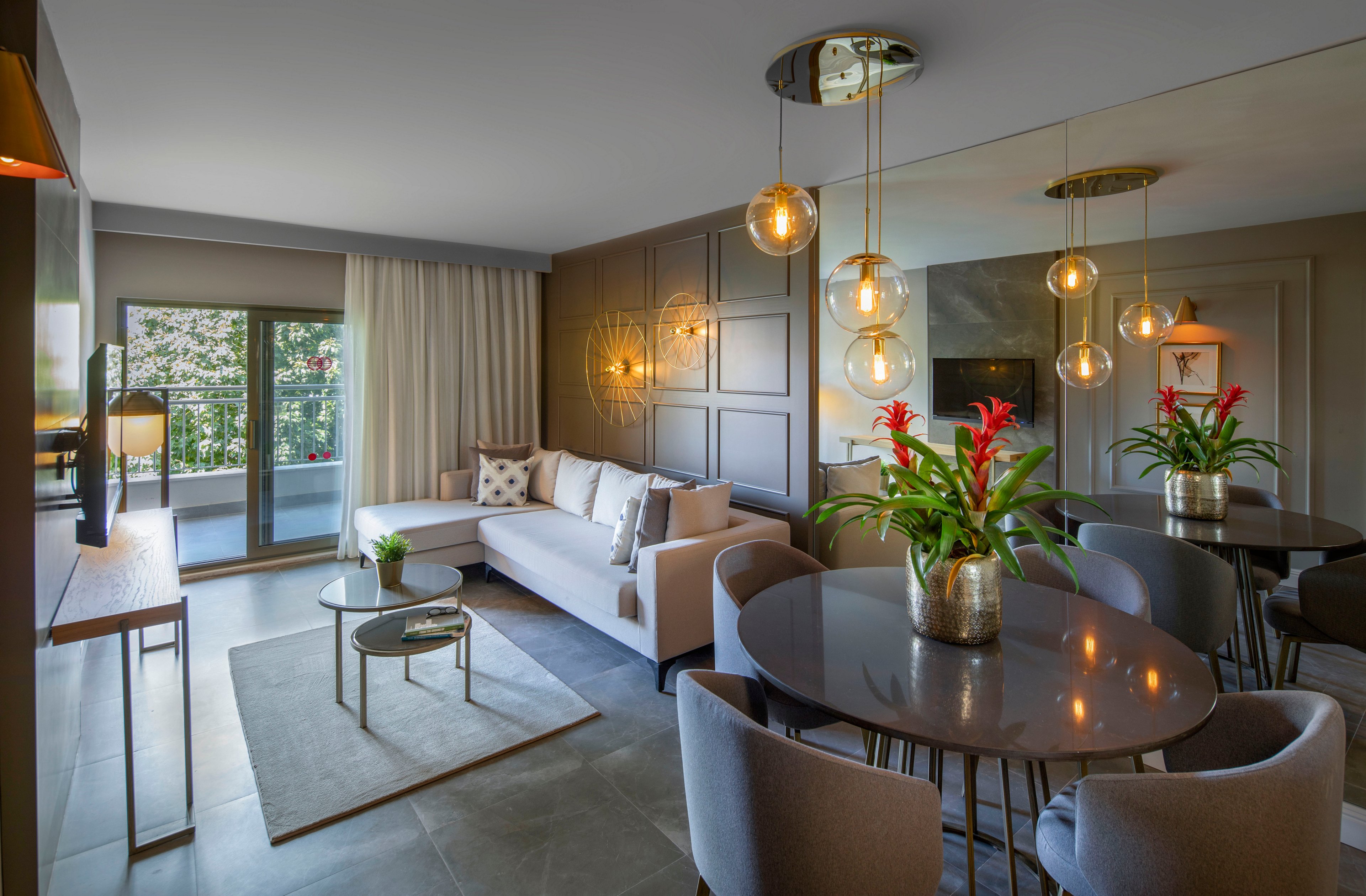 Contemporary open-plan living room features a light-colored sofa and a round dining table with upholstered chairs. Warm pendant lights hang above the table and seating area, creating a cozy atmosphere. Large windows and glass doors open to a balcony with greenery outside, while neutral walls and curtains enhance the spacious feel. Decorative plants and wall art add color and texture to the sophisticated interior design.